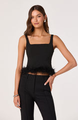 A woman is wearing a sleeveless black top with wide straps and a square neckline. The top features a fluffy feather trim along the hemline. She pairs it with high-waisted black trousers that have vertical seam details.