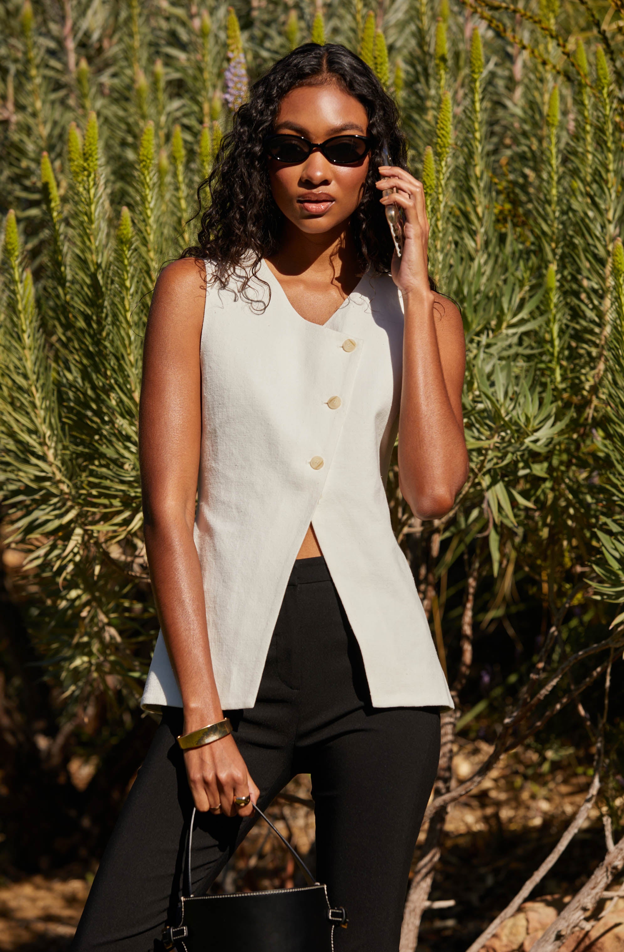 A woman is wearing a sleeveless, off-white, button-front top with a high neckline, paired with black pants. She has on dark sunglasses and is accessorizing with a gold bracelet and ring, and is holding a black handbag. She is standing outdoors in front of green, spiky plants.