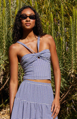 A woman is wearing a blue and white gingham two-piece outfit, consisting of a fitted crop top with a knot at the center and double spaghetti straps, paired with a matching high-waisted skirt with a gathered, ruffled hem. She is also wearing dark sunglasses and has long, curly black hair. The background features tall green plants with purple flowers.