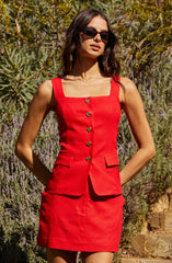 A woman is wearing a sleeveless, fitted red dress with wide shoulder straps. The dress features a square neckline, a row of decorative buttons down the front, and two small flap pockets on the front. She is also wearing large, dark sunglasses.