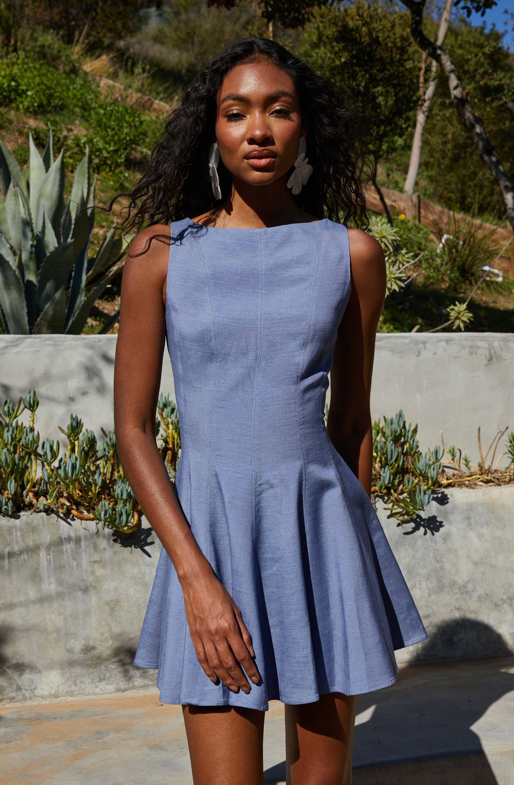 A woman wearing a sleeveless, light blue dress with a fitted bodice and a flared, pleated skirt. The dress has a simple, elegant design with clean lines. She is accessorized with large, white floral earrings.