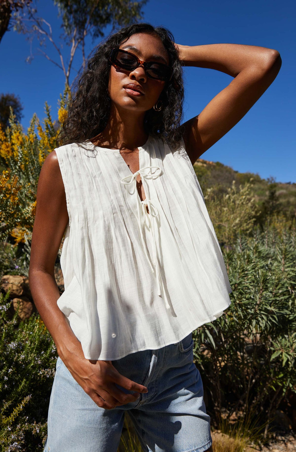 A woman is wearing a sleeveless, white, lightweight top with vertical pleats and a tie-front neckline. She pairs it with light blue denim shorts and accessorizes with dark sunglasses.