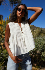 A woman is wearing a sleeveless, white, lightweight top with vertical pleats and a tie-front neckline. She pairs it with light blue denim shorts and accessorizes with dark sunglasses.