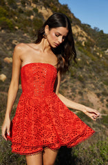 A woman is wearing a strapless, knee-length dress made of bright red fabric with intricate eyelet embroidery. The dress features a fitted bodice and a flared skirt with scalloped edges at the hem. The embroidery includes circular and floral patterns throughout the dress.