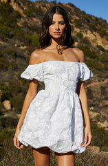 A woman is wearing an off-the-shoulder white dress with puffed short sleeves and a floral textured pattern throughout. The dress has a fitted bodice with a gathered waist and a slightly puffed, layered skirt that ends above the knees.