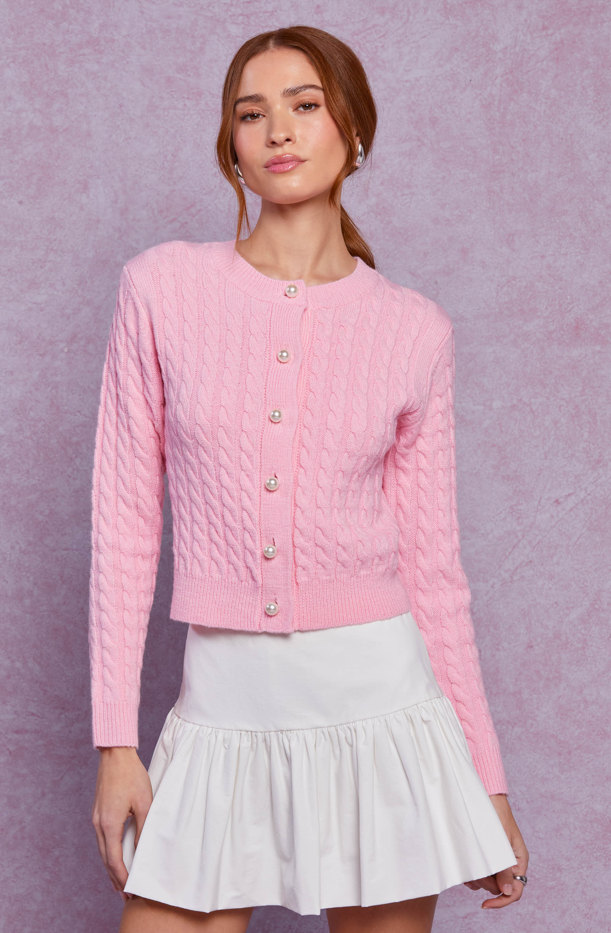 A woman is wearing a light pink, cable-knit cardigan with pearl buttons down the front. The cardigan has a round neckline and long sleeves. She pairs it with a high-waisted, white, ruffled skirt.