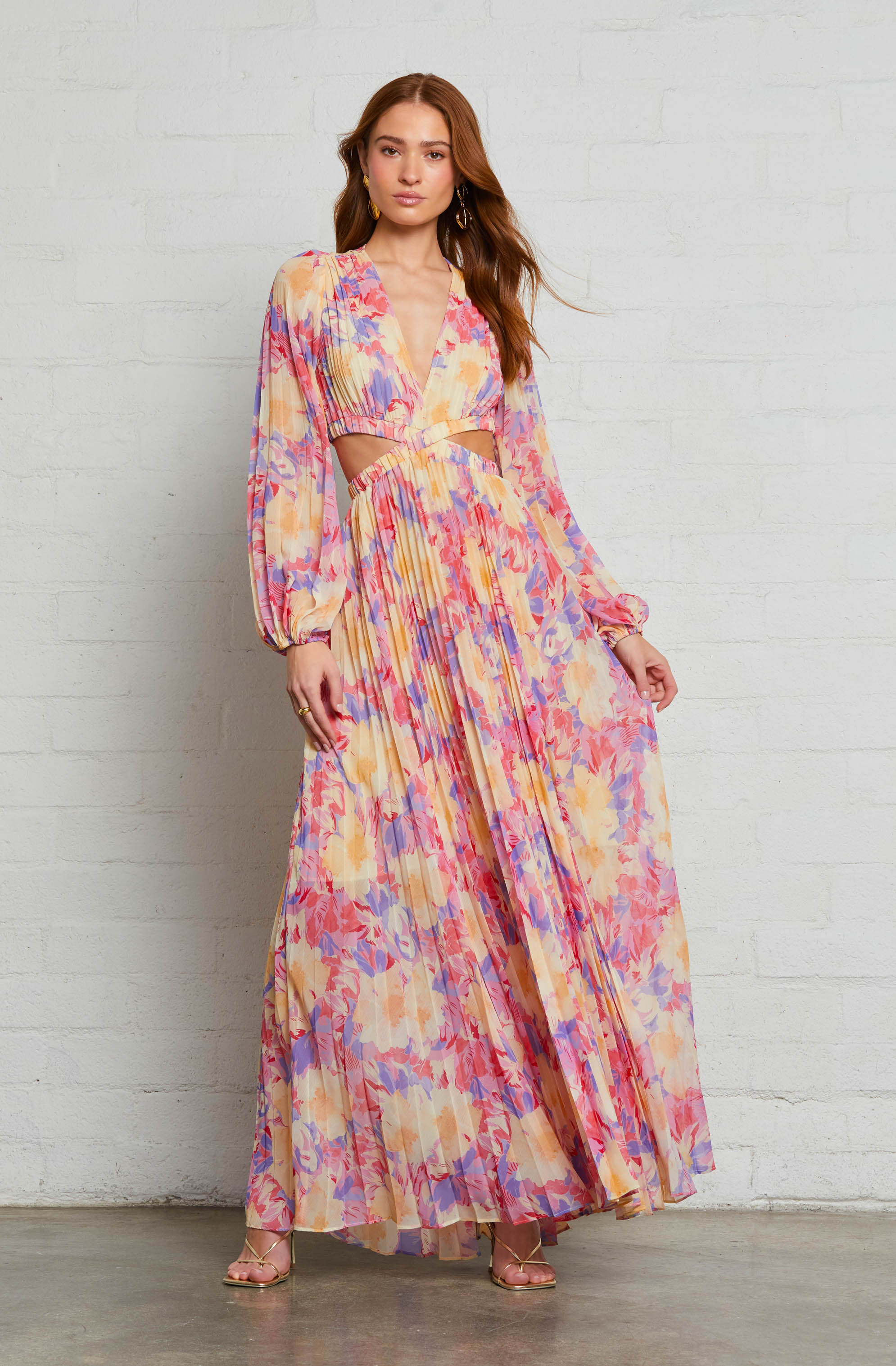 A woman is wearing a long, flowy dress with a colorful, abstract floral pattern in shades of pink, purple, yellow, and orange. The dress features a deep V-neckline with a wrap-style front, cut-out details at the waist, and long, billowy sleeves gathered at the wrists. She is also wearing open-toe heels and gold hoop earrings.