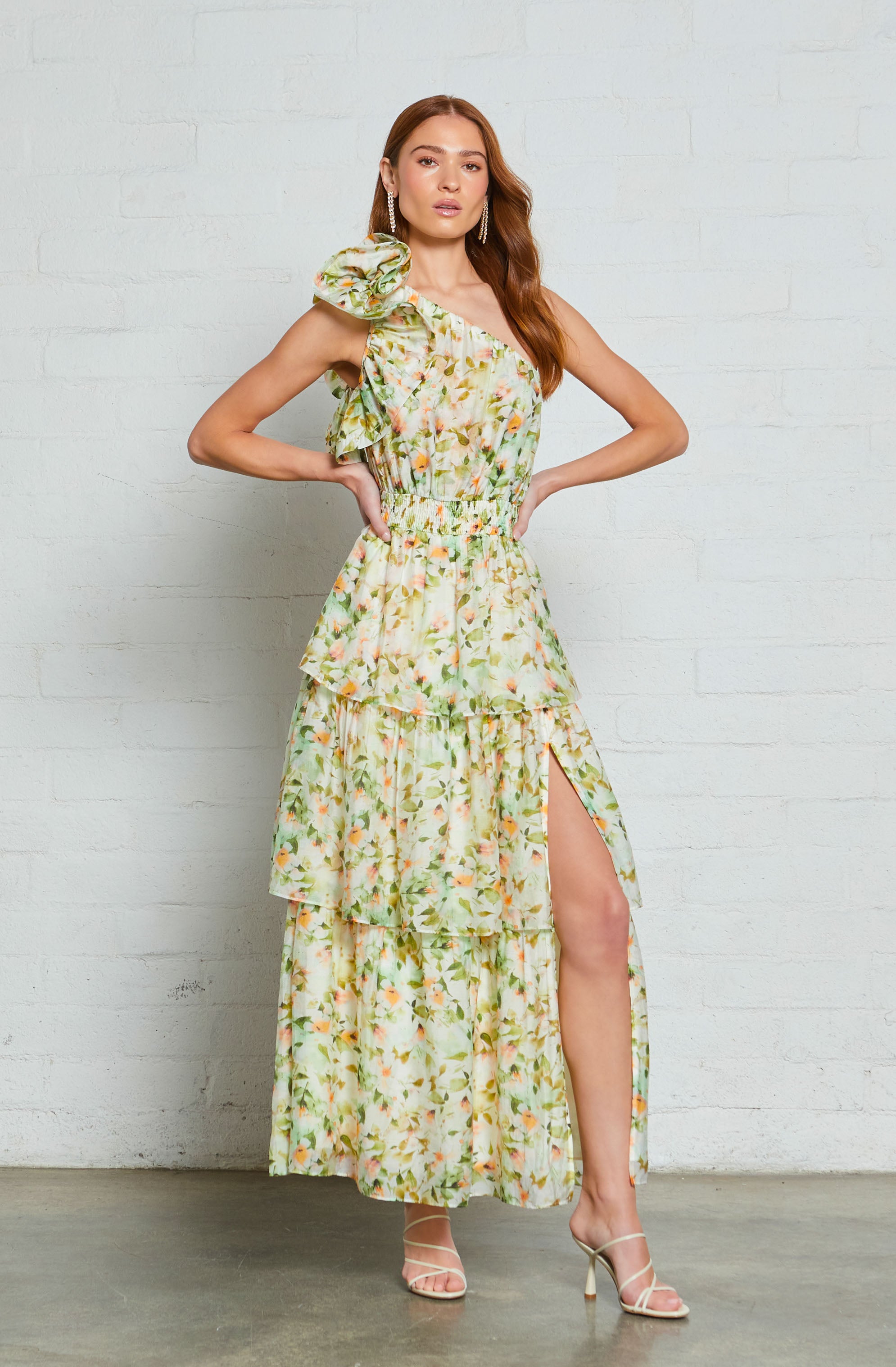 A woman is wearing a long, sleeveless dress with a pastel floral pattern. The dress features ruffled details on the shoulder and a slit on one side, revealing her leg. She is also wearing strappy high heels.