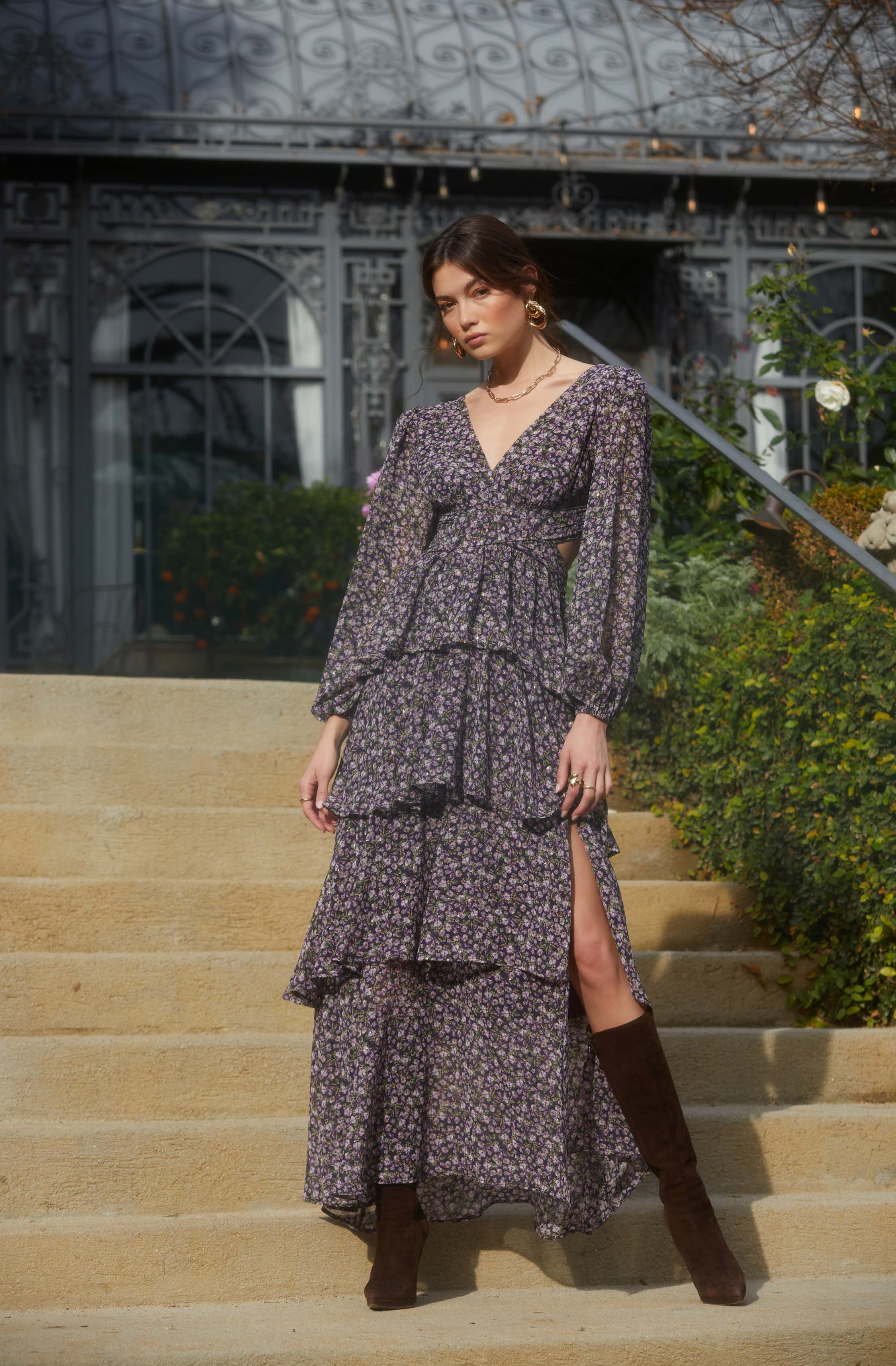 A woman is wearing a long, dark-colored dress with a small floral pattern. The dress features a deep V-neckline, long sheer sleeves, and a fitted waistband. The skirt is tiered with multiple ruffles, including a high slit on one side revealing her leg and brown knee-high boots.