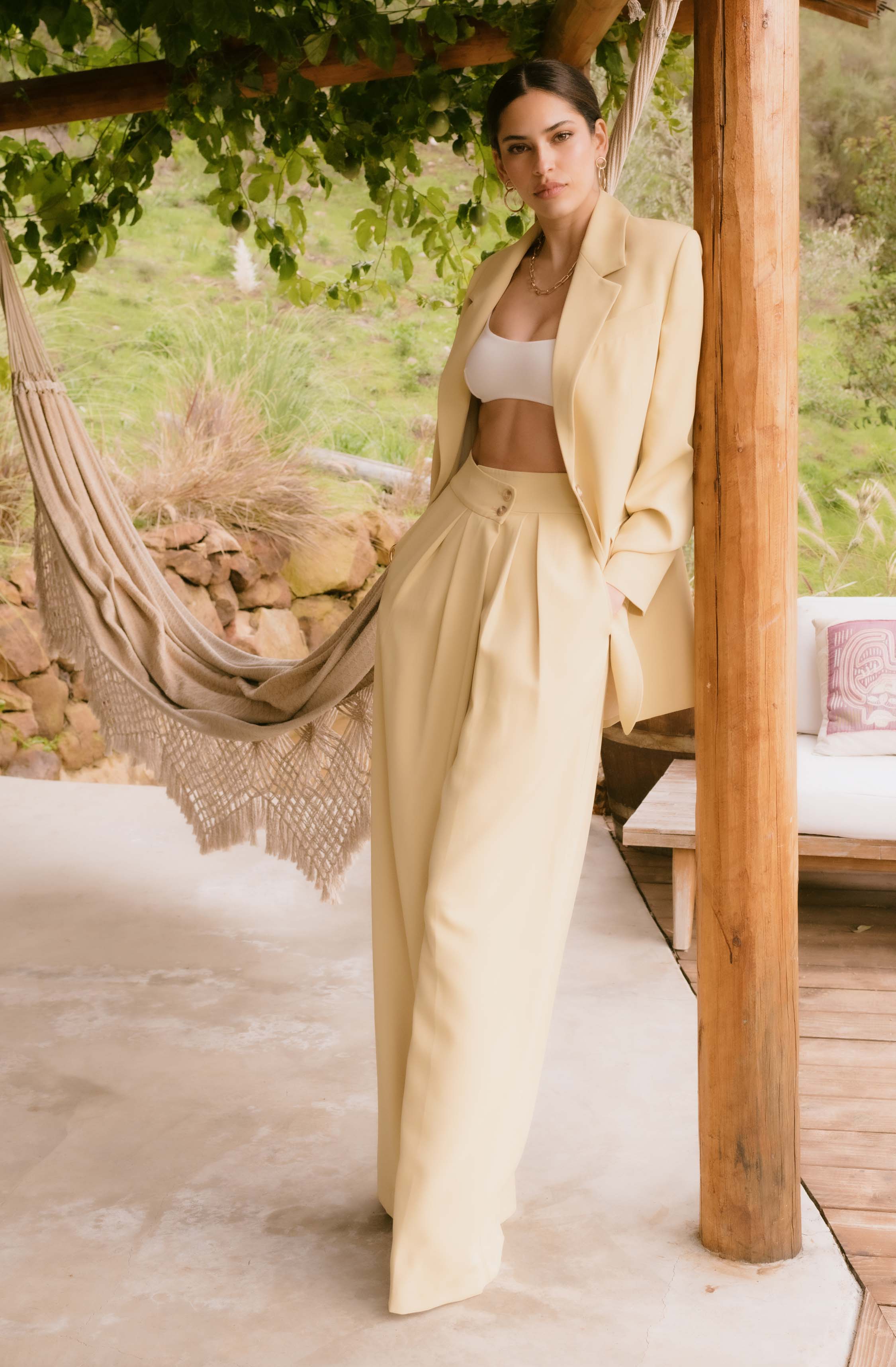 The person is wearing a beige suit with high-waisted, wide-leg trousers and a matching blazer. Underneath, they have on a white crop top. They are also accessorized with large hoop earrings and a gold chain necklace.