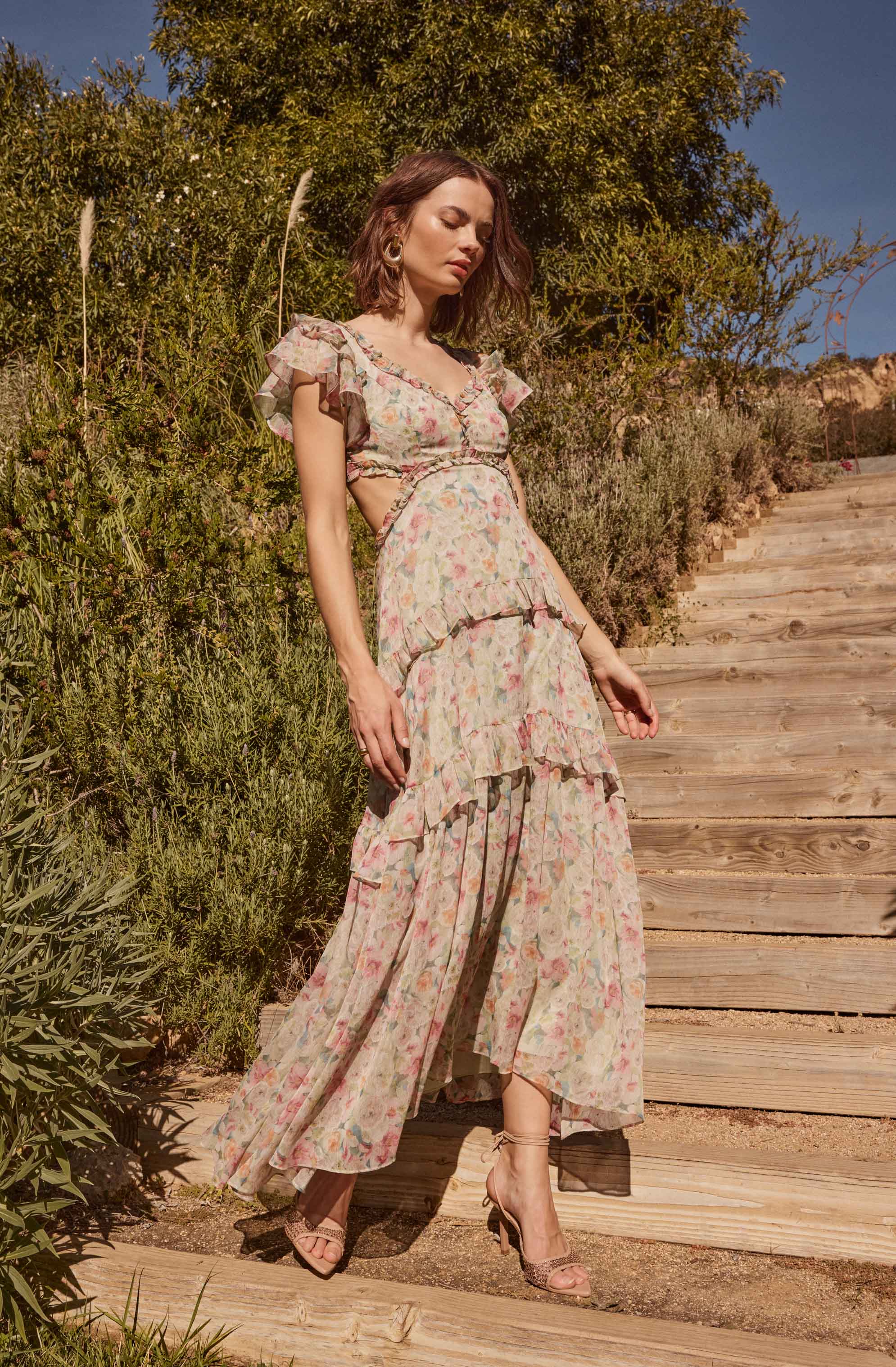 A woman is wearing a flowy, pastel-colored floral dress with ruffled short sleeves, a fitted bodice, and a layered, tiered skirt. The dress features soft, muted tones with a delicate floral pattern, and she pairs it with beige high-heeled sandals.