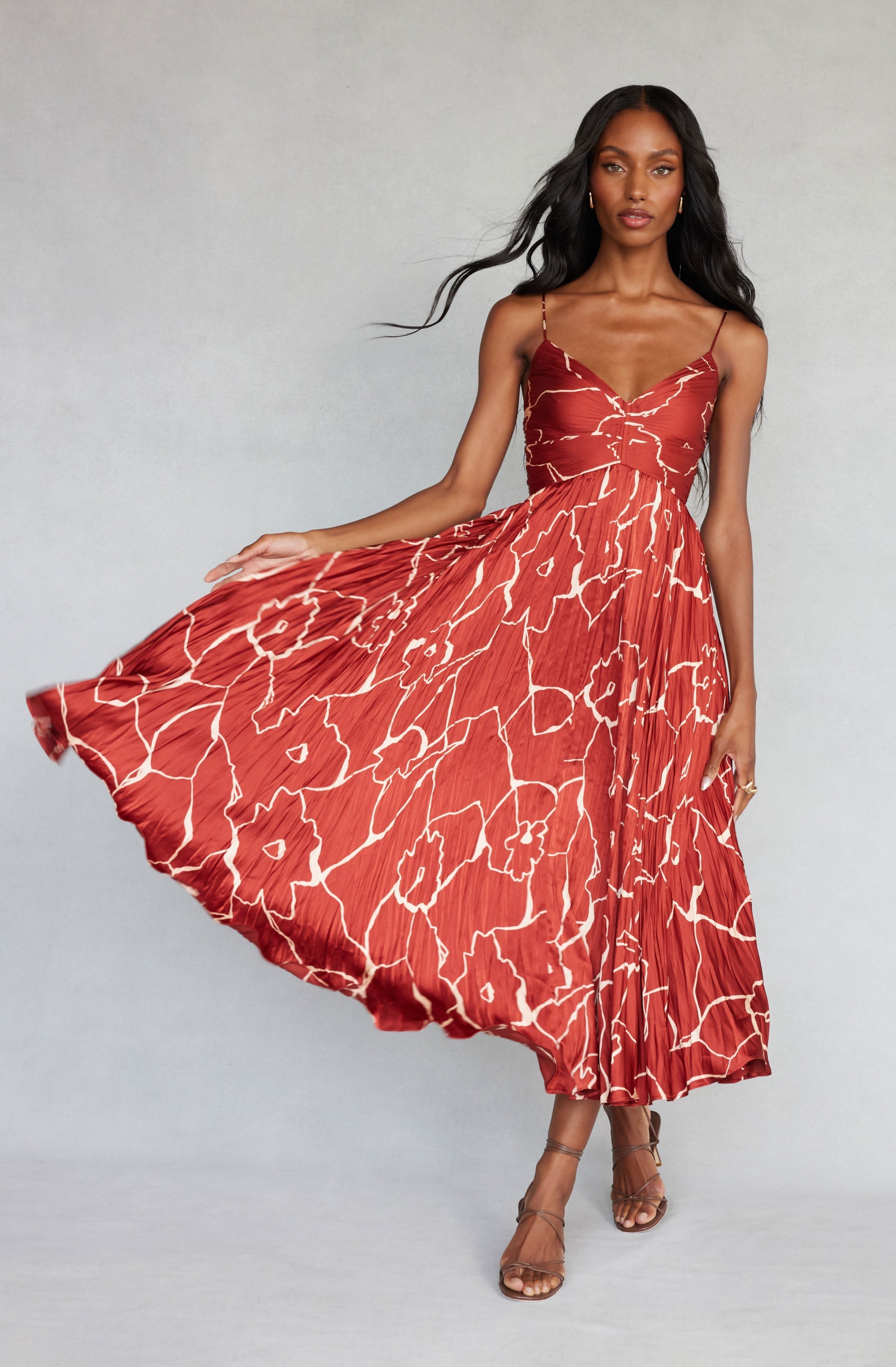 A woman wearing a red, sleeveless, slip dress with thin spaghetti straps. The dress features a floral pattern with large white outlines of flowers. She is holding the side of the dress outwards with one hand, creating a flowing effect. She has long, wavy black hair and is wearing nude high-heeled sandals.