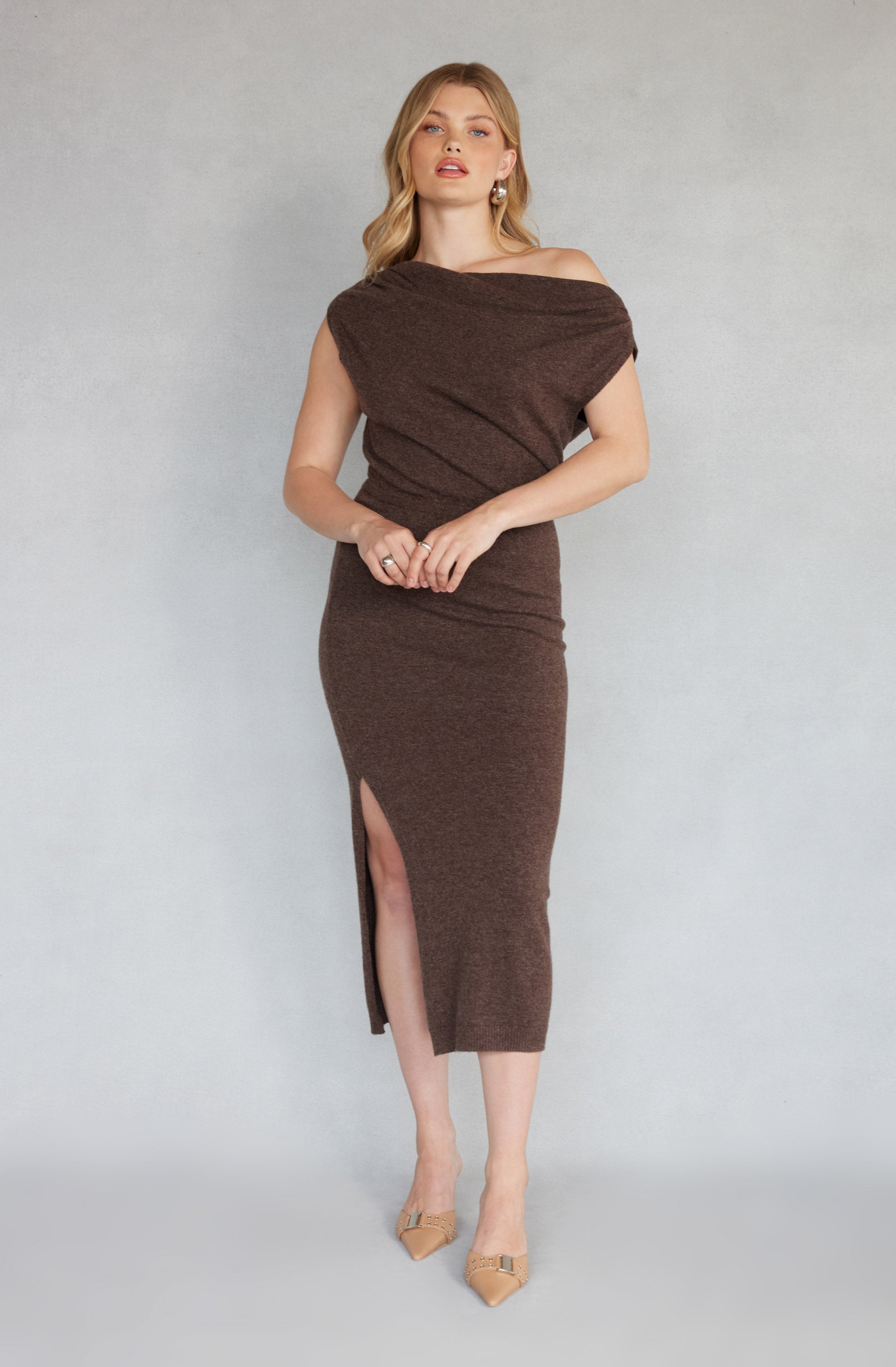 A woman is wearing a dark brown, off-the-shoulder knit dress with a loose, draped top that creates a wide neckline. The dress is fitted at the waist and has a high slit on one side, reaching below the knees. She is also wearing nude-colored pointed-toe heels and gold hoop earrings.