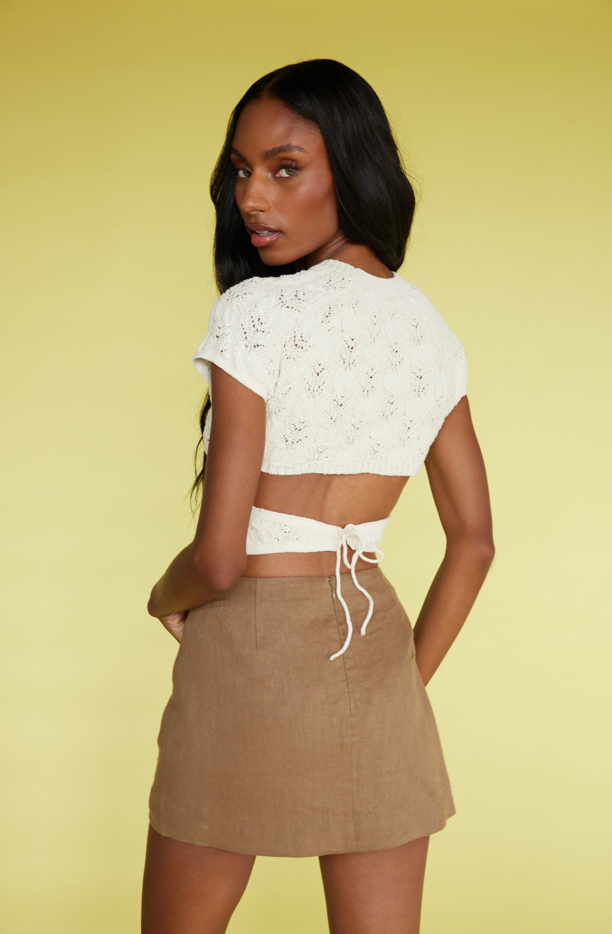 A woman is wearing a cream-colored, short-sleeved, knitted crop top with a tie at the back. She pairs it with a tan, high-waisted, A-line mini skirt made of a textured fabric.