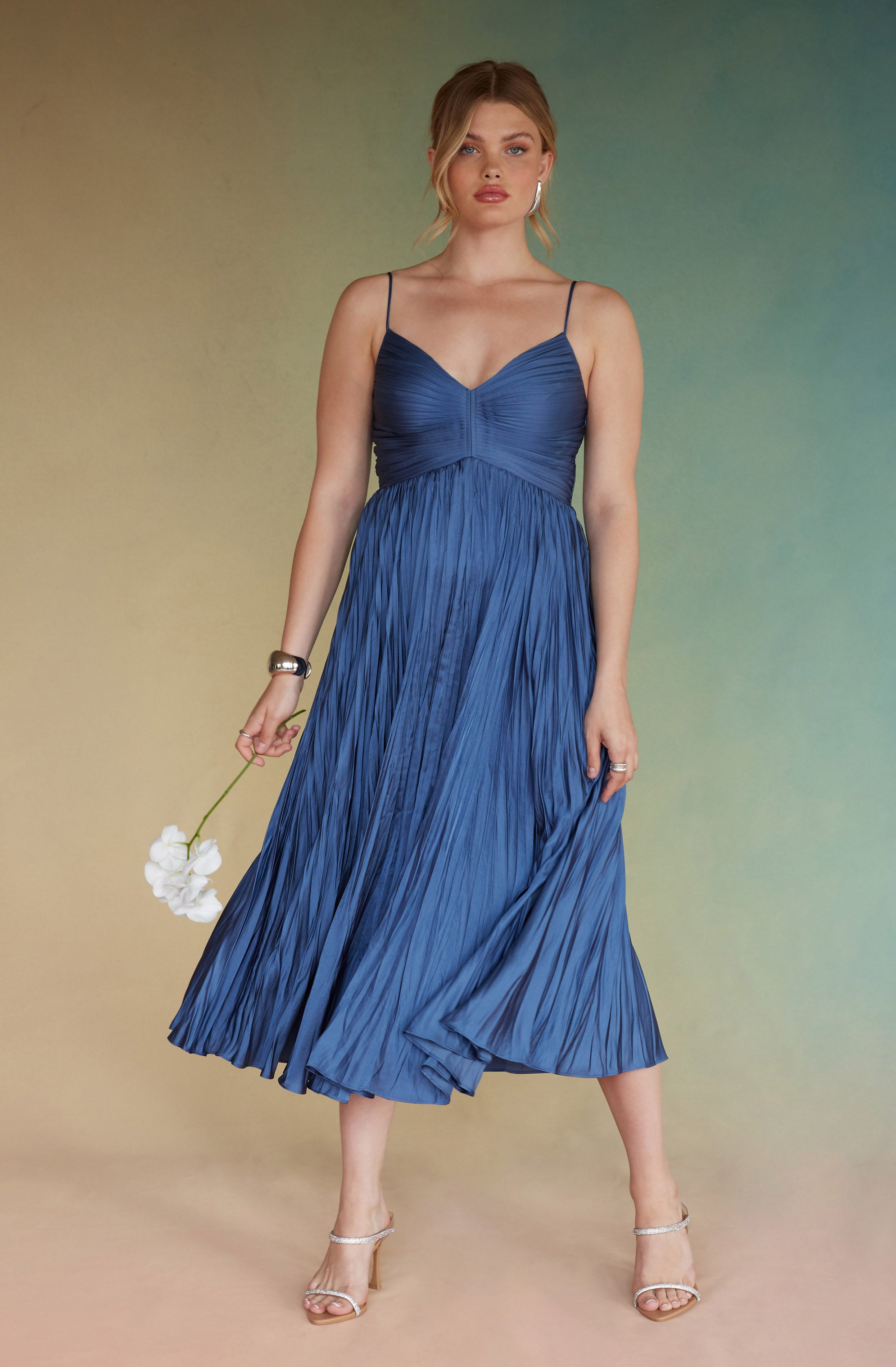A woman is wearing a sleeveless, deep blue dress with thin spaghetti straps. The dress has a ruched bodice with a V-neckline and a flowing, pleated skirt that reaches mid-calf. She pairs the dress with light-colored, open-toed heels and accessorizes with a silver bracelet on her left wrist and a ring on her right hand.