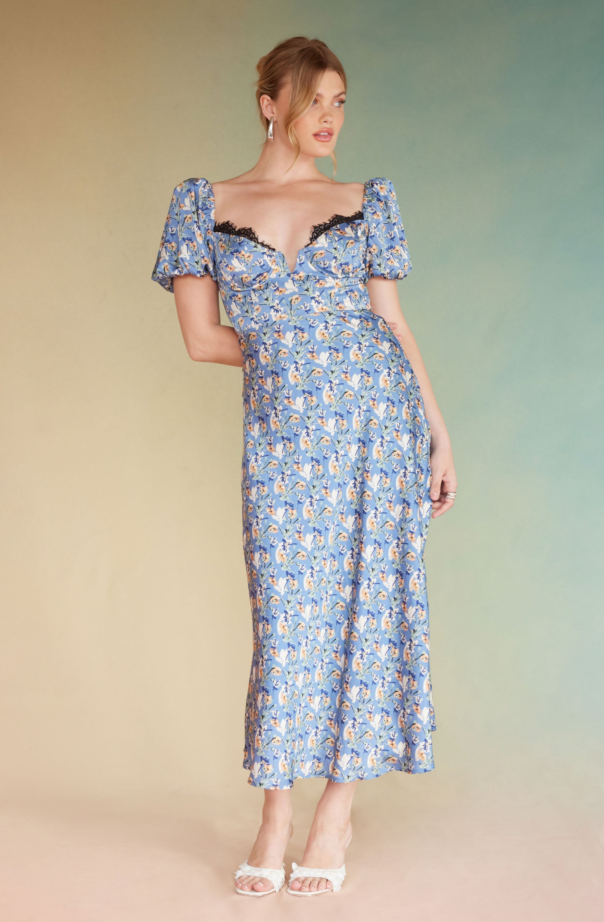 A woman is wearing a floor-length, blue floral dress with puffed short sleeves. The dress features a sweetheart neckline accented with black lace, and the fabric has a pattern of small, multicolored flowers. She is also wearing white high-heeled sandals with open toes.
