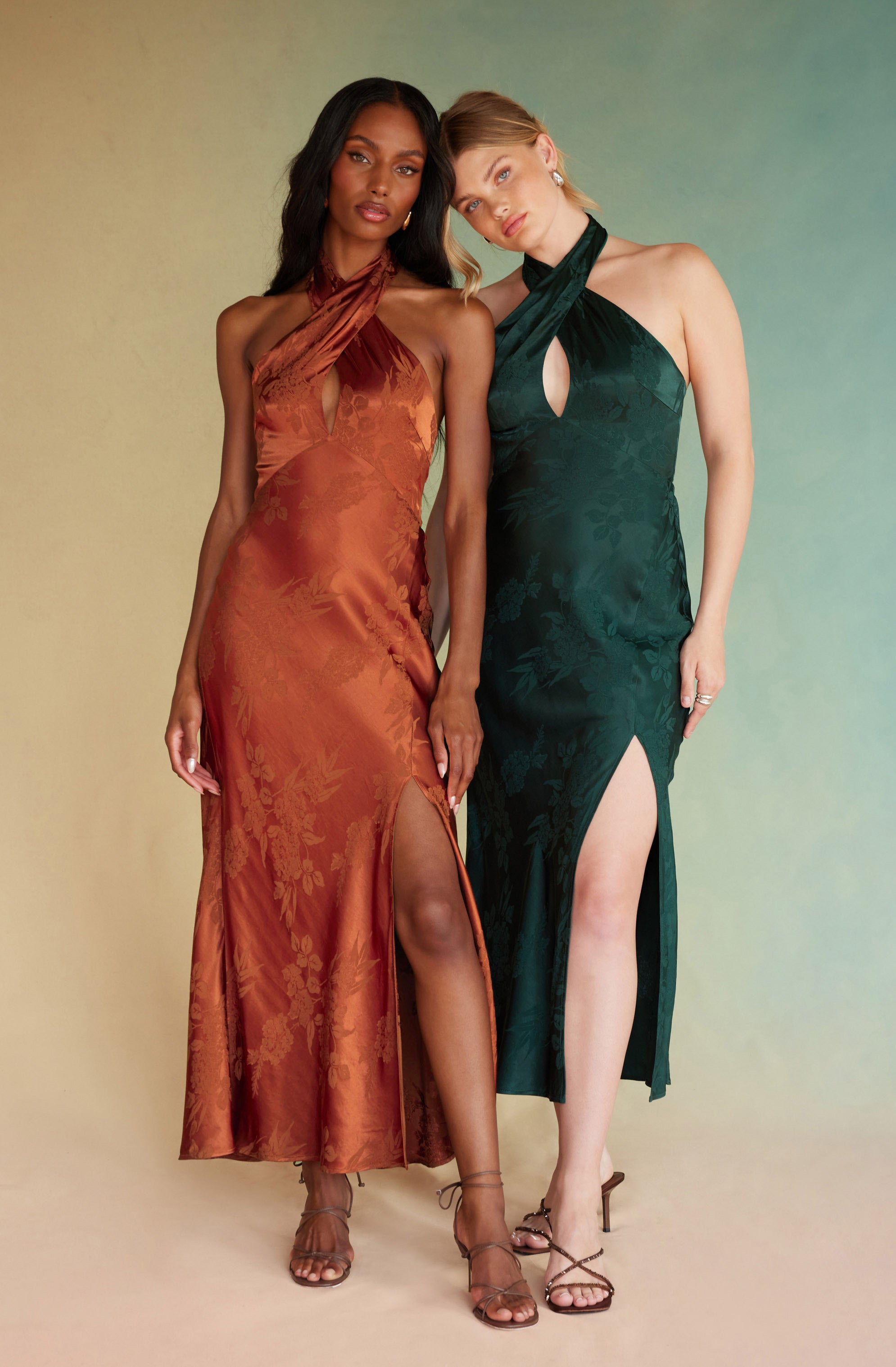 Two women are wearing elegant, sleeveless, floor-length satin dresses with high slits. The woman on the left is dressed in a rust-colored gown with a halter neckline that wraps around her neck, and the dress has a subtle floral pattern. The woman on the right is dressed in a deep emerald green gown with a halter neckline that has a keyhole cutout in the front and a similar floral pattern. Both women are wearing strappy high-heeled sandals suitable for formal occasions.