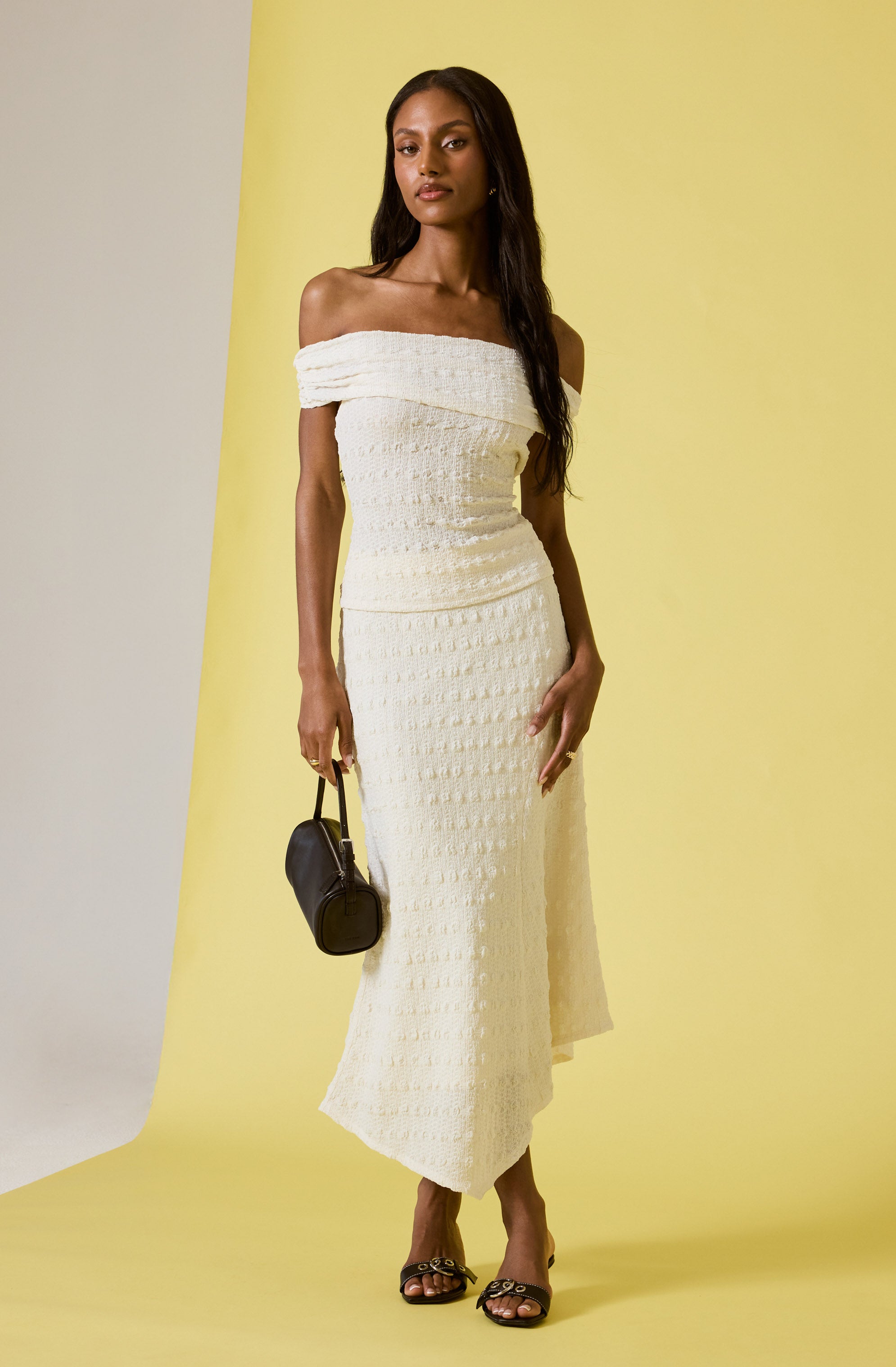 A woman is wearing a white off-the-shoulder maxi dress with a textured, woven pattern. She is holding a small black handbag in her right hand and is wearing strappy black and gold heeled sandals.