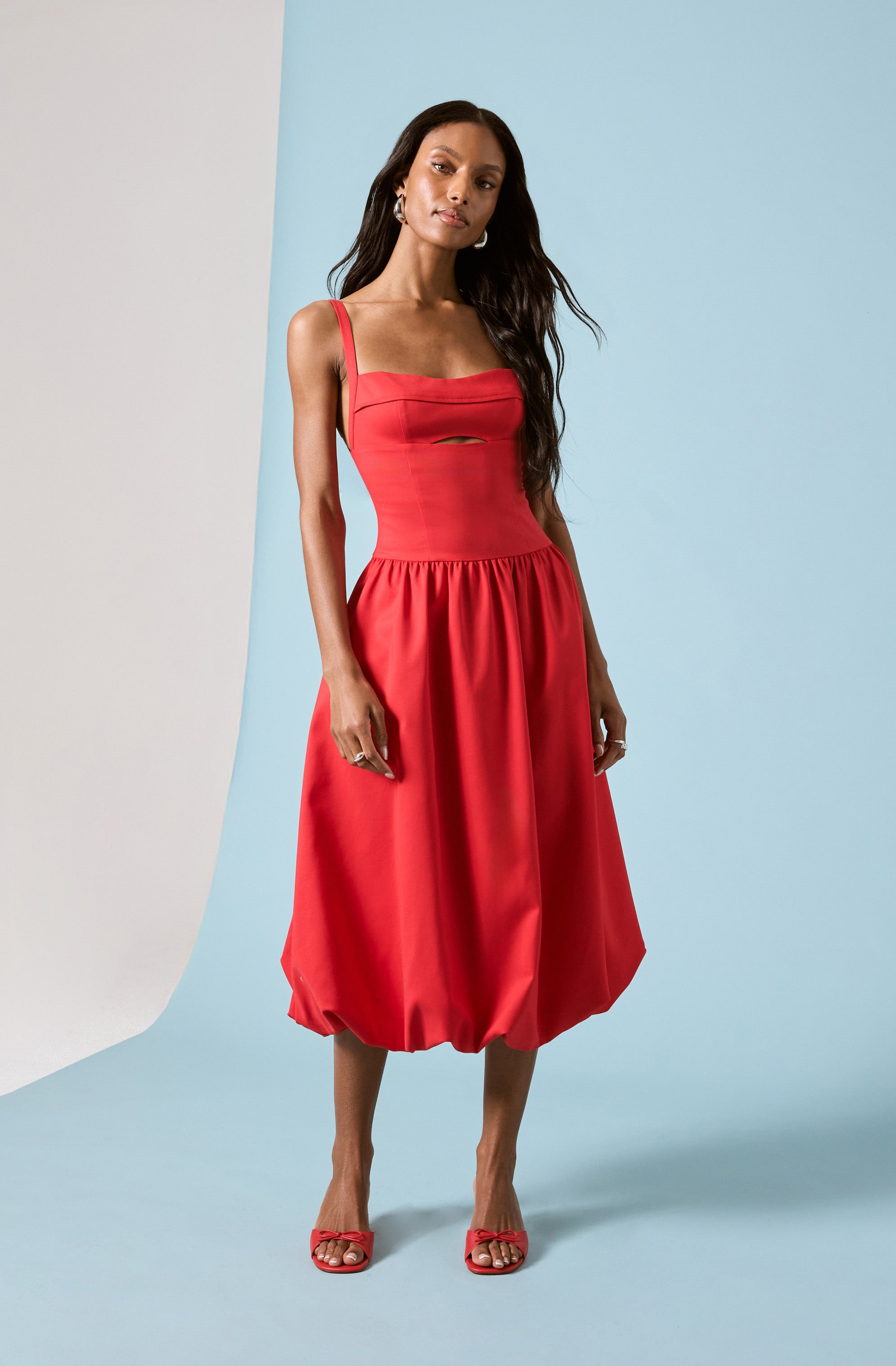 A woman is wearing a vibrant red dress with thin shoulder straps and a fitted bodice featuring a small cutout detail at the chest. The skirt of the dress is gathered and has a voluminous, balloon-like shape that extends to mid-calf length. She completes her outfit with matching red heeled sandals.