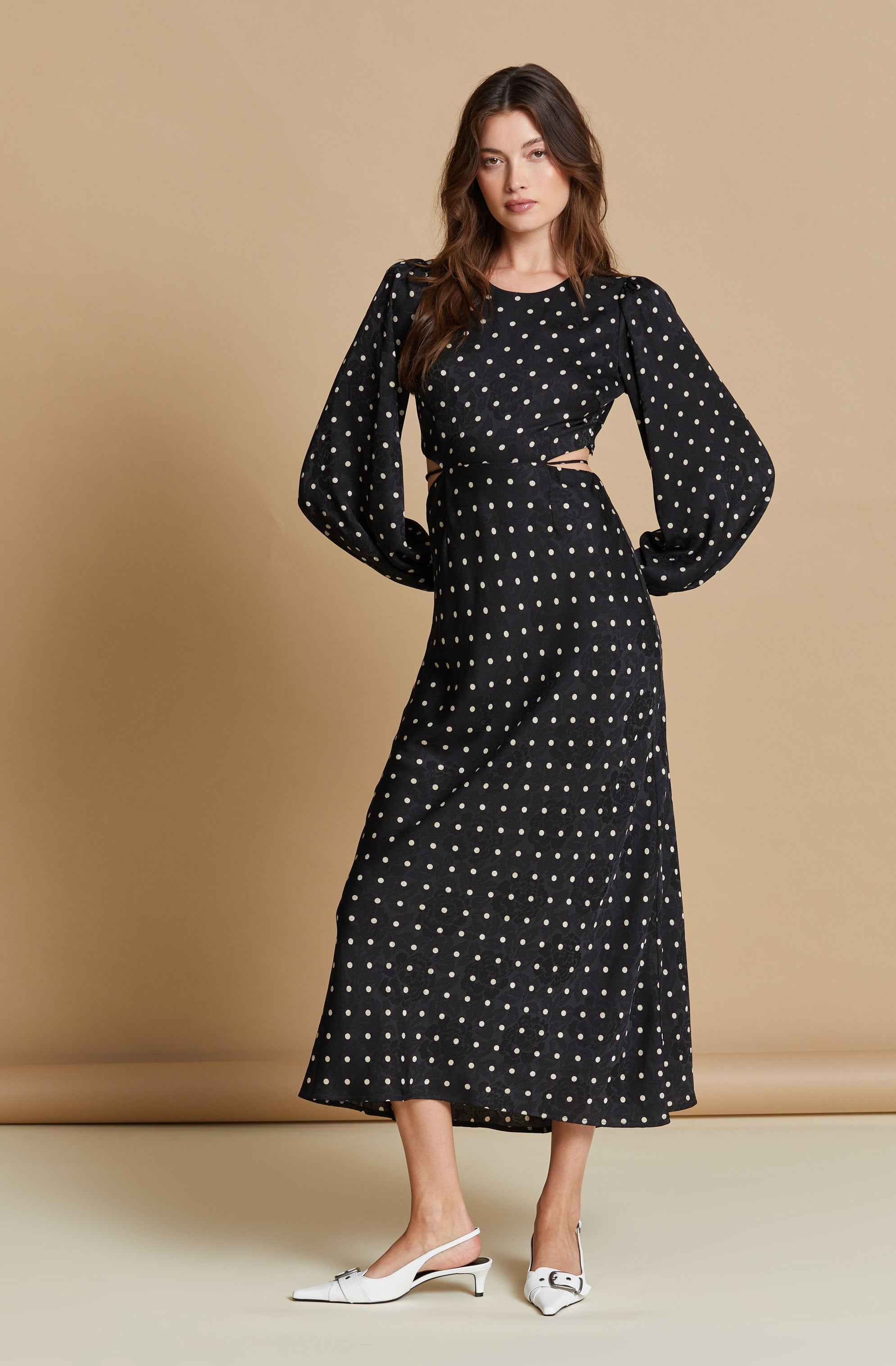 A woman is wearing a black dress with white polka dots, featuring puffed long sleeves and cut-out details at the waist. She pairs the dress with white pointed-toe heels that have a small heel and a decorative buckle.