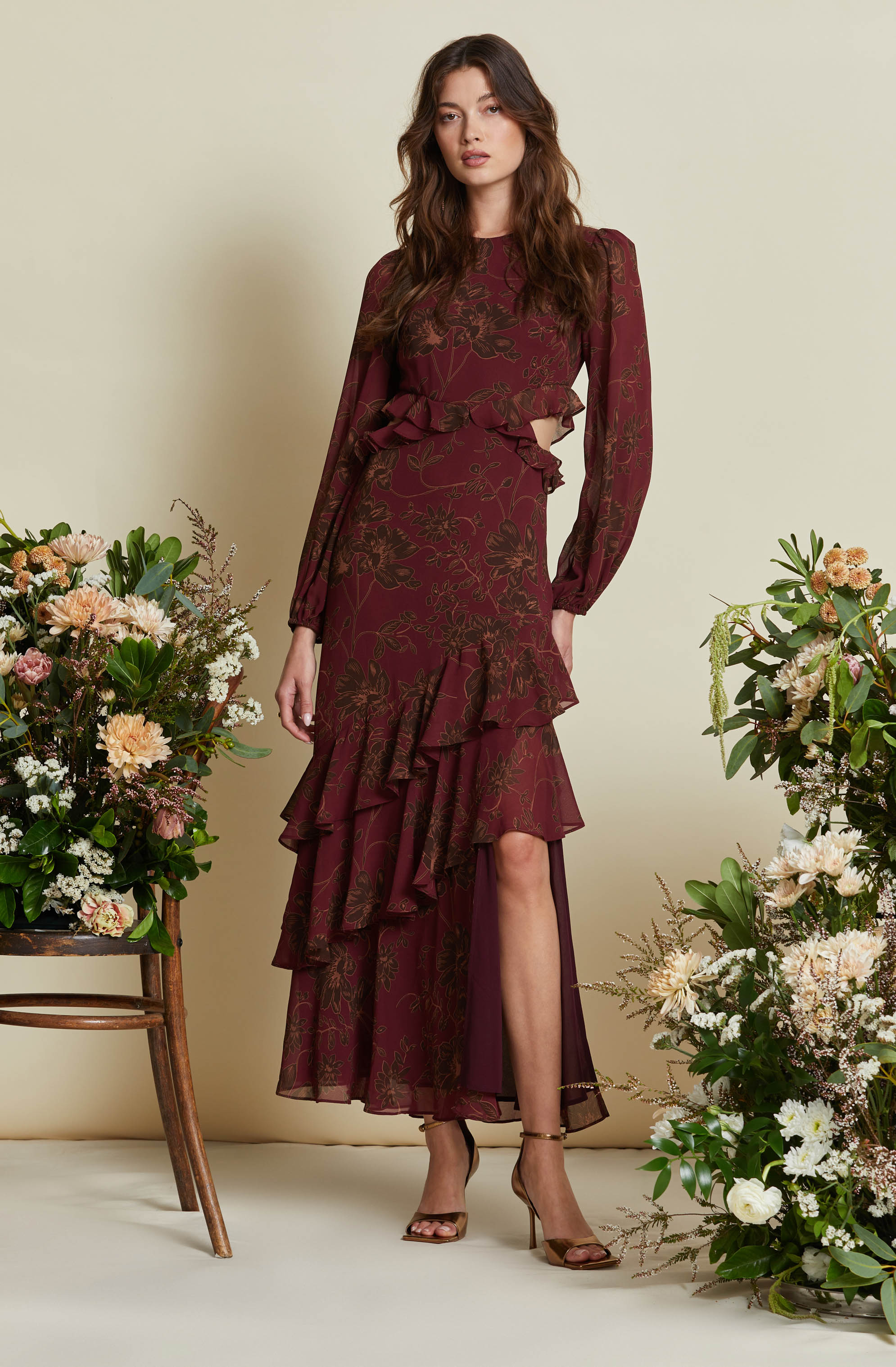 A woman is wearing a long, flowy, burgundy dress with a floral pattern. The dress has long sleeves and a ruffled hem that cascades asymmetrically down the front, with a high slit revealing her leg. She is also wearing nude-colored high-heeled sandals.