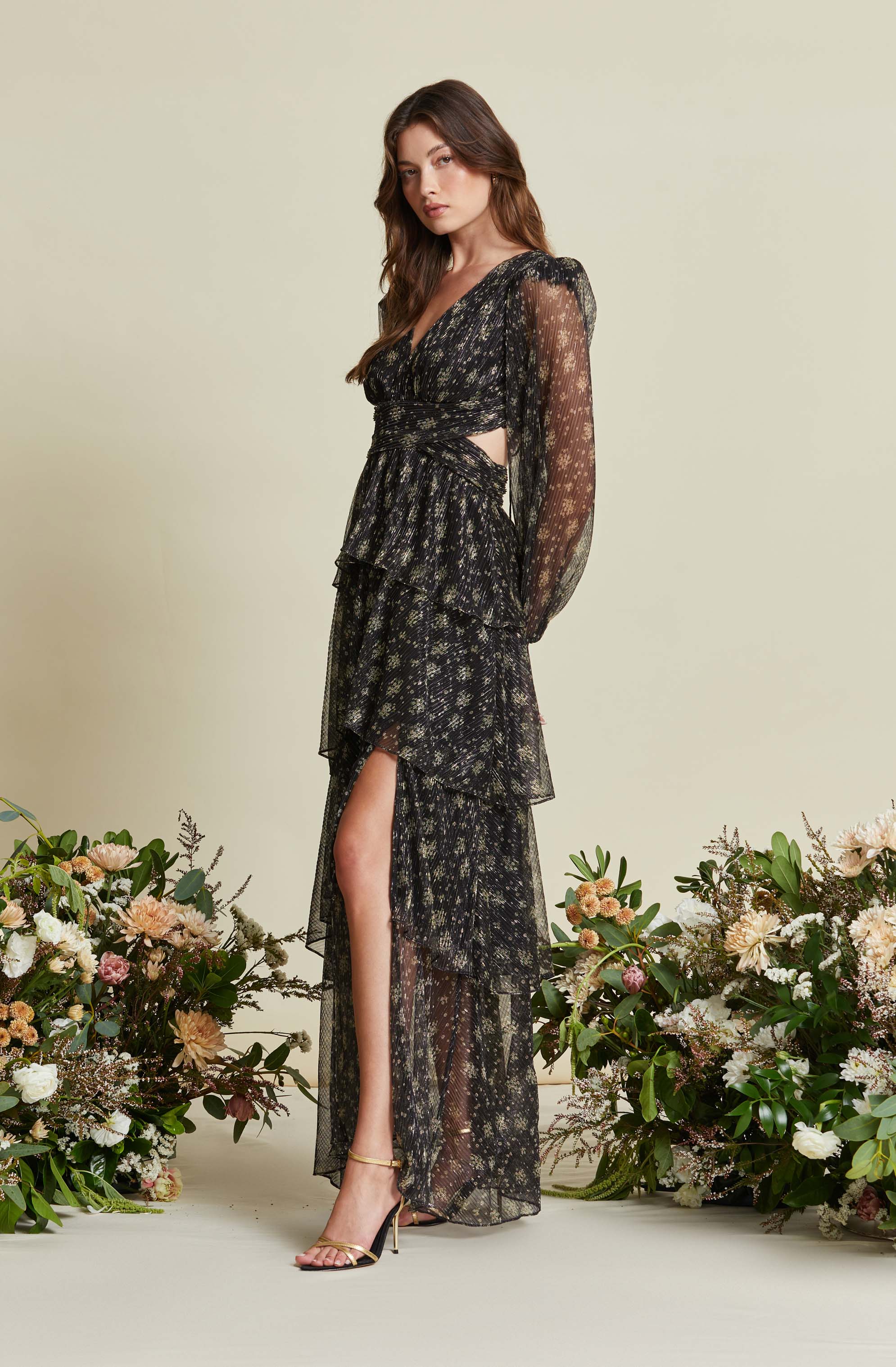 A woman is wearing a black, sheer, long-sleeved dress with a floral pattern in shades of gold and beige. The dress features a plunging V-neckline, cut-out sides at the waist, tiered ruffles on the skirt with a high slit revealing her leg, and ankle-strap high heels. The background is light-colored, and she is surrounded by floral arrangements with various flowers and greenery.