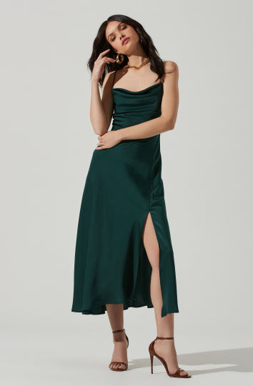 A woman is wearing a dark emerald green satin dress with thin spaghetti straps. The dress has a fitted bodice with a draped neckline and a high slit on the left side that reveals her leg. She accessorizes with large gold hoop earrings, a choker necklace, and tan high-heeled sandals.