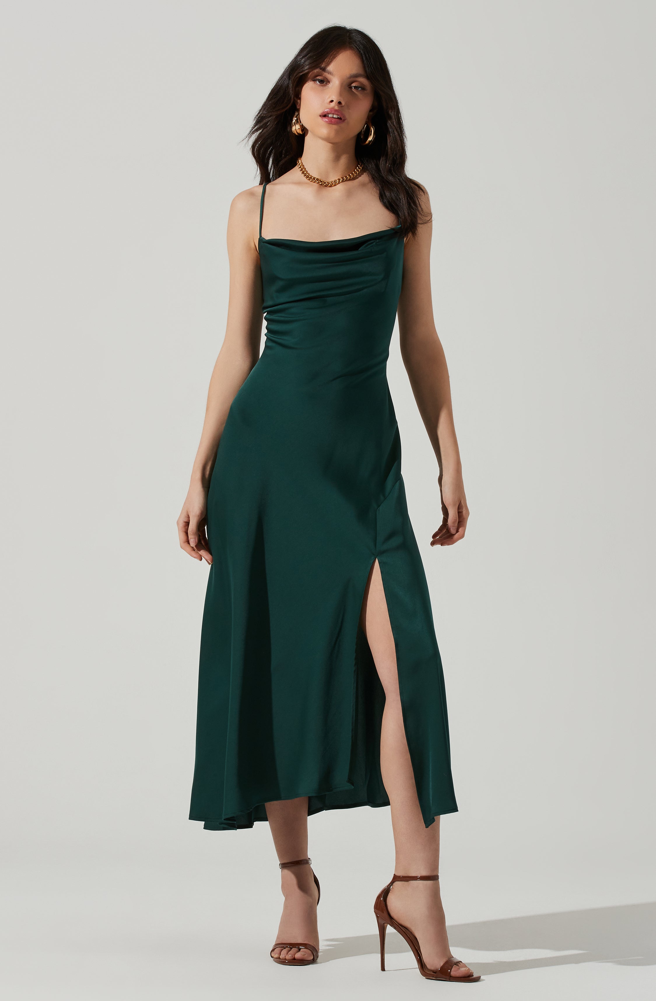 A woman is wearing a dark green satin slip dress with spaghetti straps, featuring a cowl neckline and a high slit on the front left side. She accessorizes with large gold hoop earrings and a chunky gold chain necklace. She is also wearing tan high-heeled sandals with ankle straps.