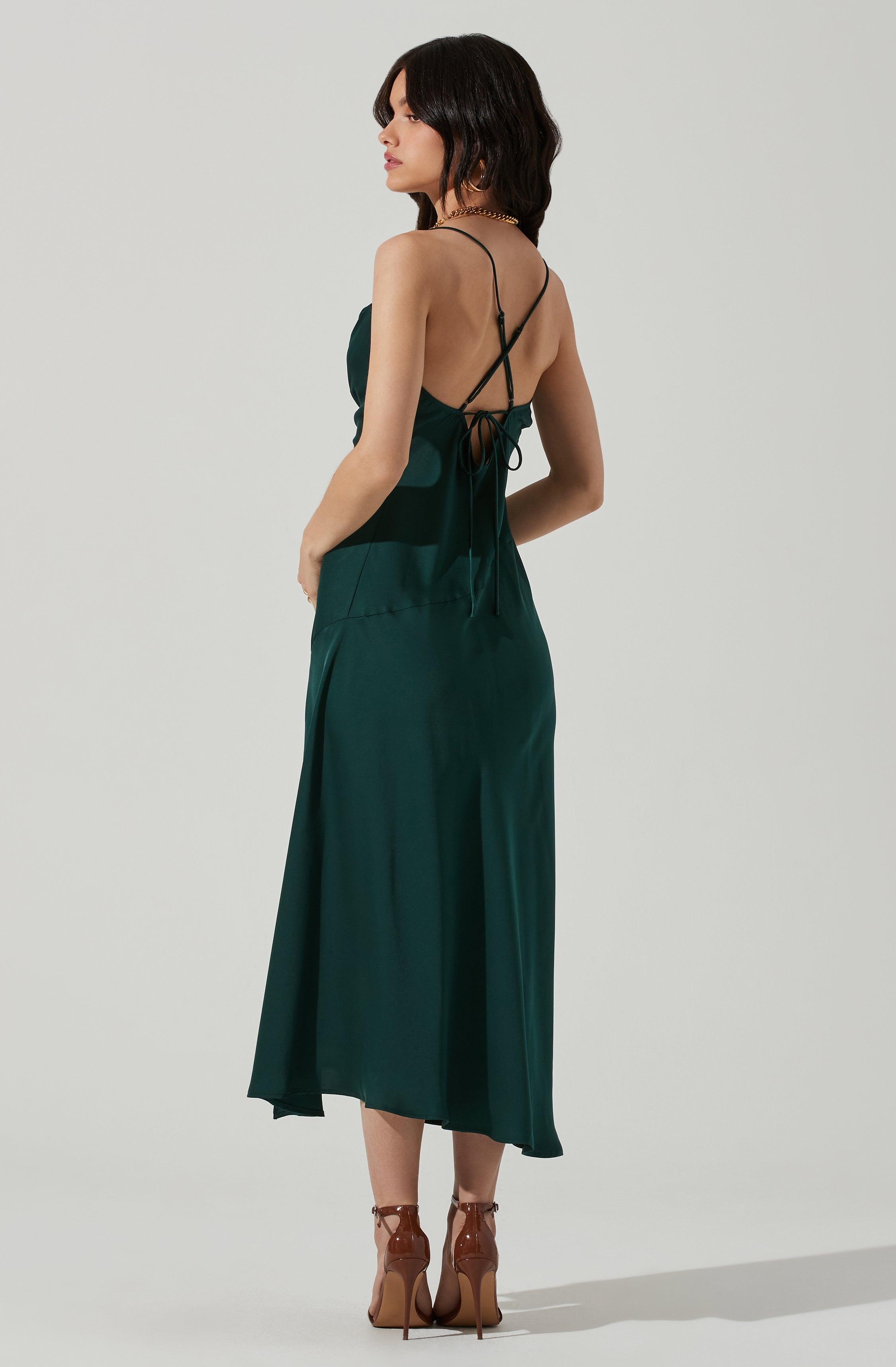 A woman is wearing a dark green satin slip dress with thin spaghetti straps that crisscross and tie at the back. The dress has a flowy, ankle-length hem. She is also wearing brown high-heeled sandals with ankle straps. The accessories include a chunky gold chain necklace and hoop earrings. Her dark hair is styled in loose waves.