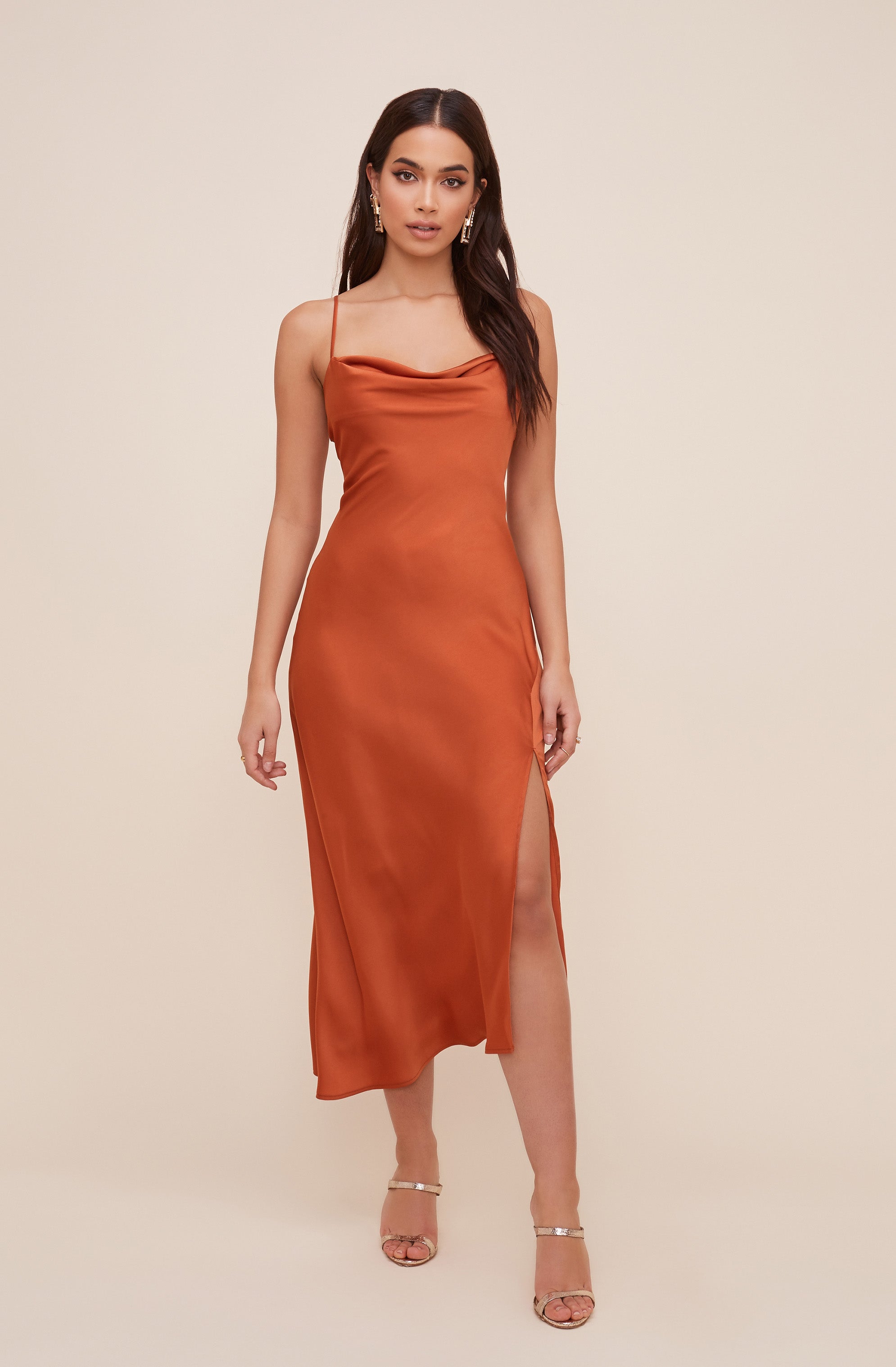 A woman is wearing a burnt orange satin slip dress with thin spaghetti straps and a draped cowl neckline. The dress features a high slit on one side, revealing her leg. She has paired it with strappy, open-toed heels. She is also wearing gold earrings.