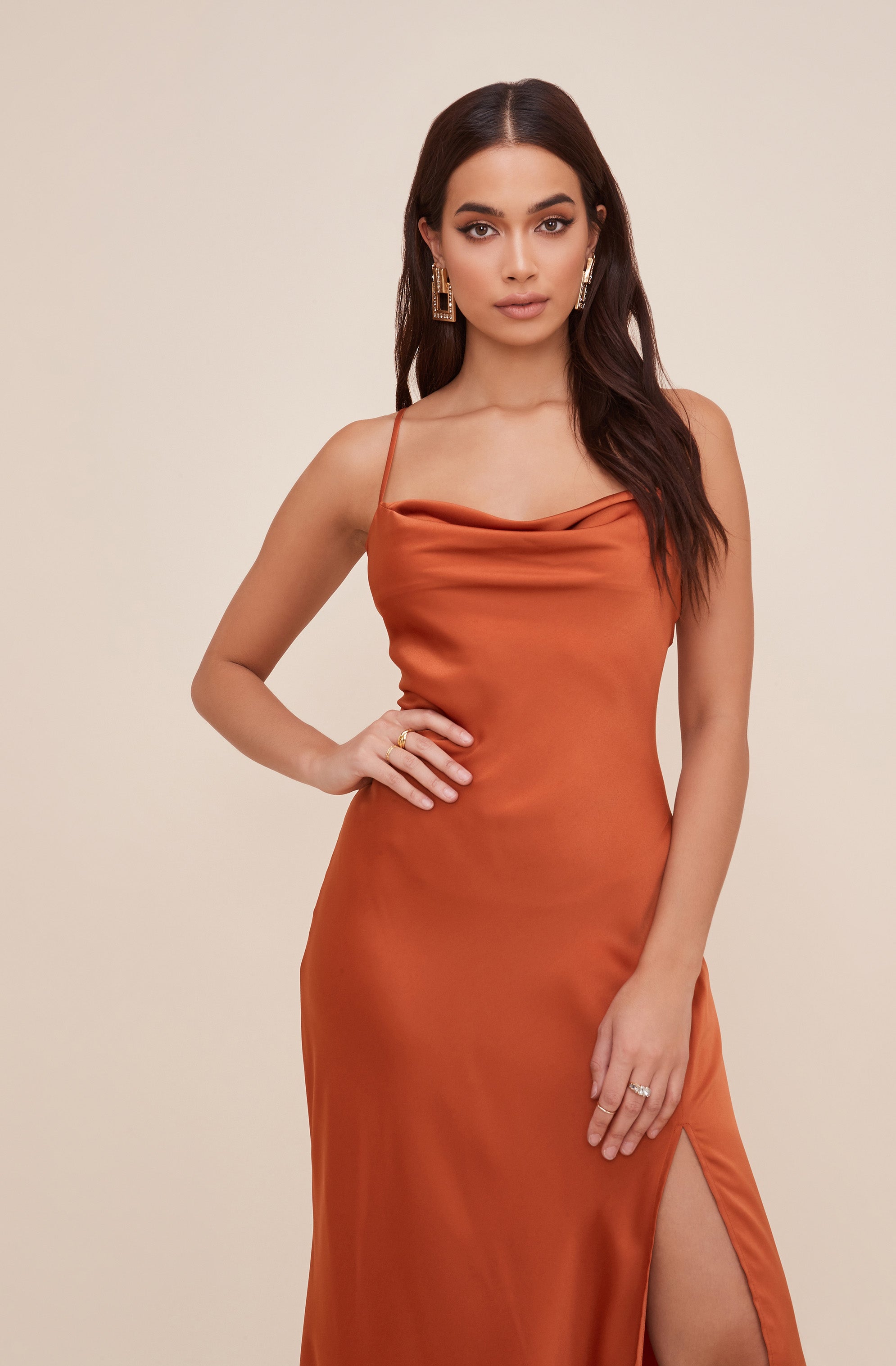 A woman is wearing a satin, rust-colored slip dress with thin spaghetti straps, a cowl neckline, and a high slit on the side.