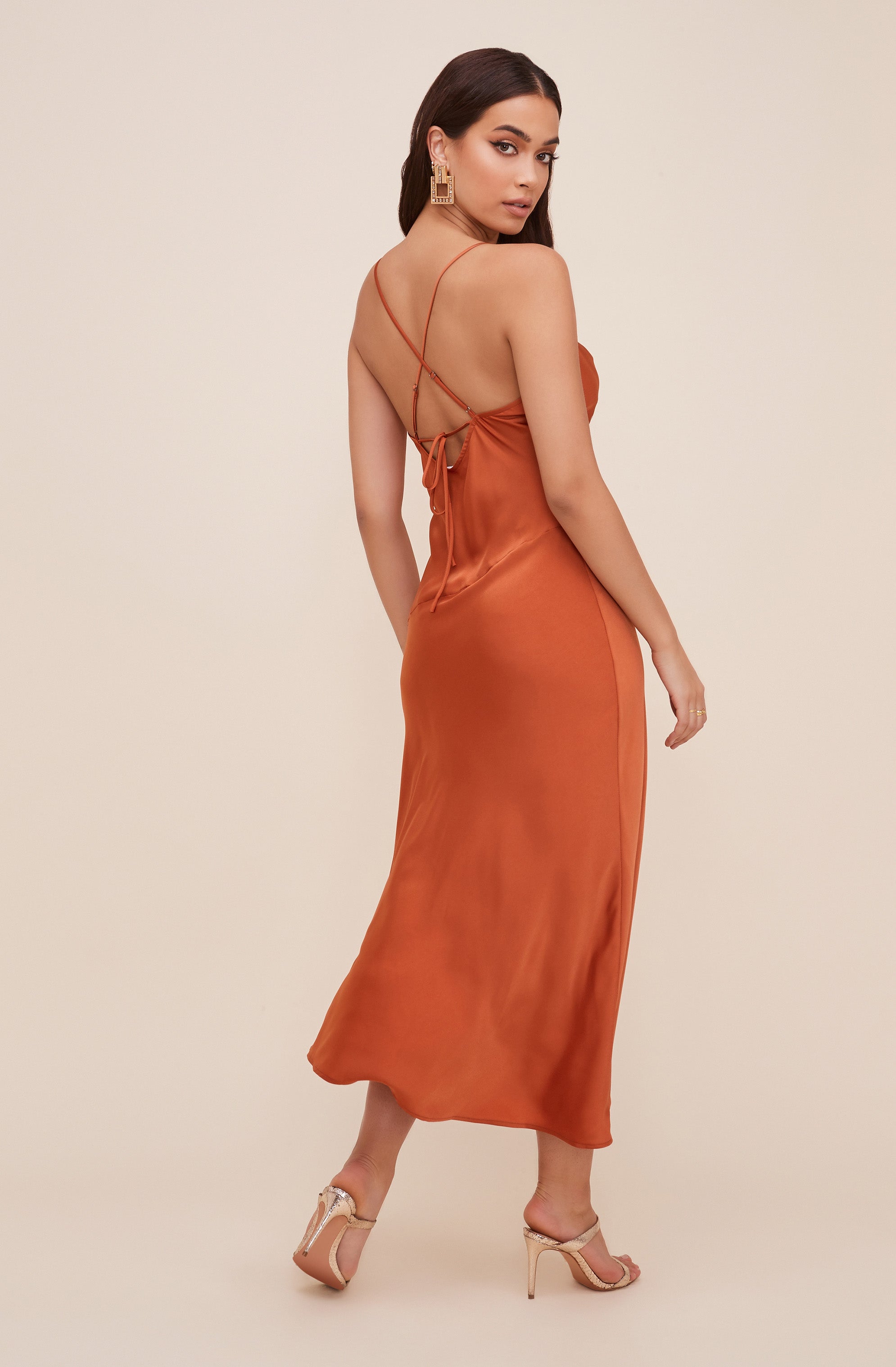 A woman is wearing a long, silky, orange satin dress with thin spaghetti straps that cross at the back and tie into a bow. She is also wearing high-heeled, open-toe shoes with ankle straps, and accessorized with large, rectangular gold earrings.