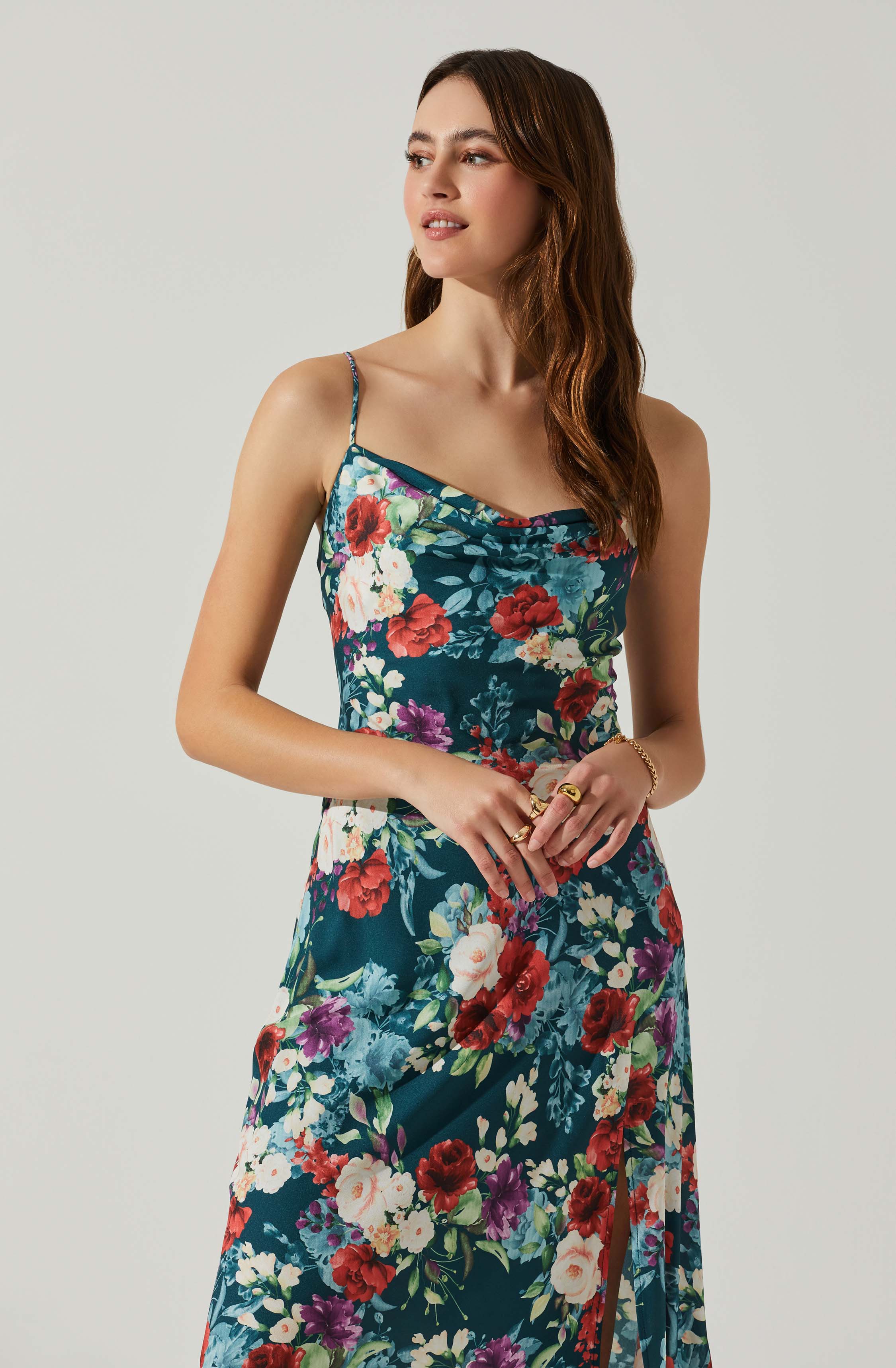 A woman is wearing a sleeveless, floral dress with thin spaghetti straps. The dress features a dark teal background adorned with a colorful pattern of red, white, purple, and pink flowers with green leaves. The fabric appears smooth and silky, draping elegantly over her figure. She is accessorized with multiple rings and a bracelet.