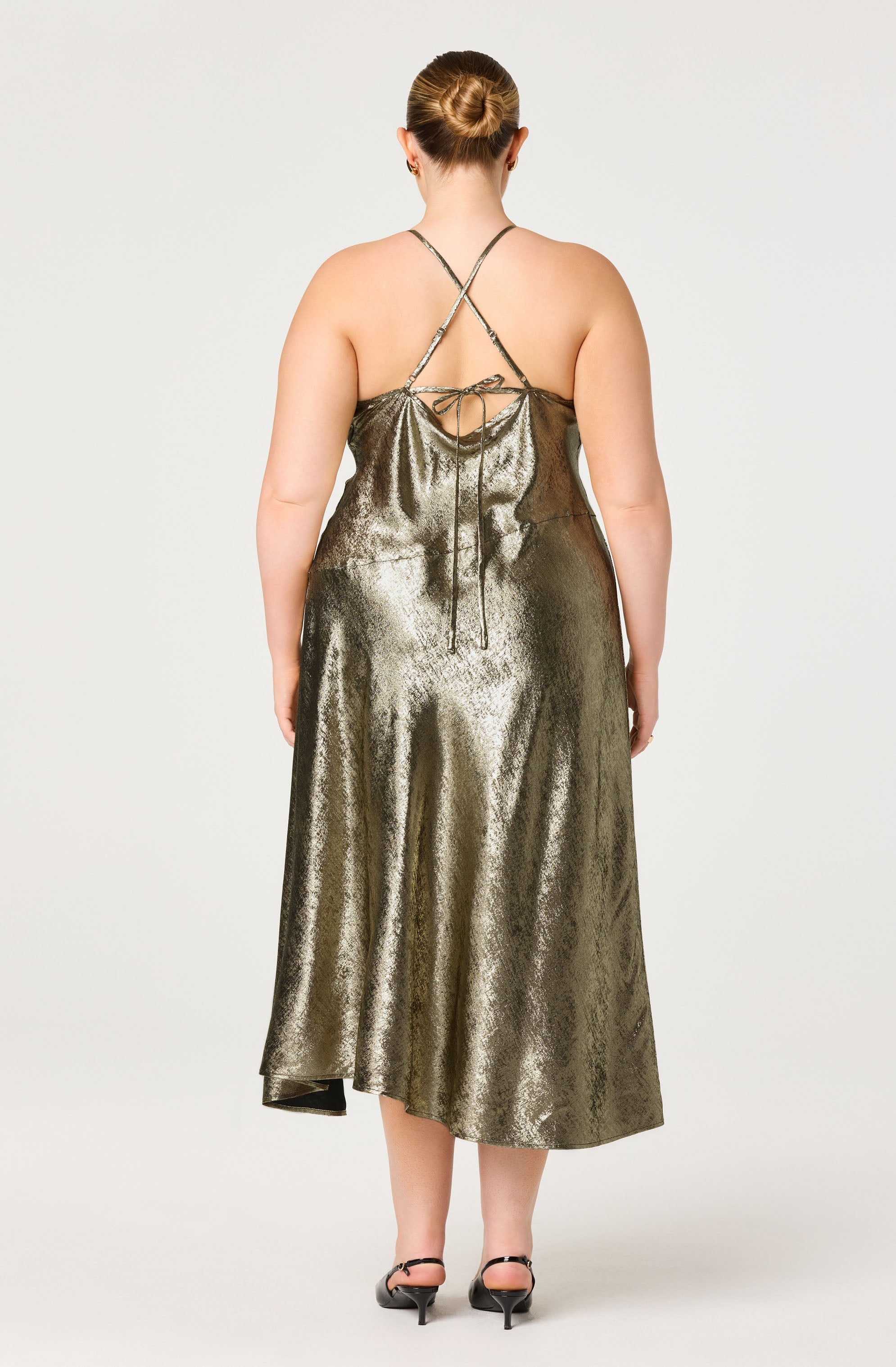 A woman is wearing a metallic gold, sleeveless, ankle-length dress with thin spaghetti straps that cross at the back and tie at the mid-back. The fabric has a shiny, textured appearance, and the dress drapes loosely around her body. She is wearing black high-heeled shoes.