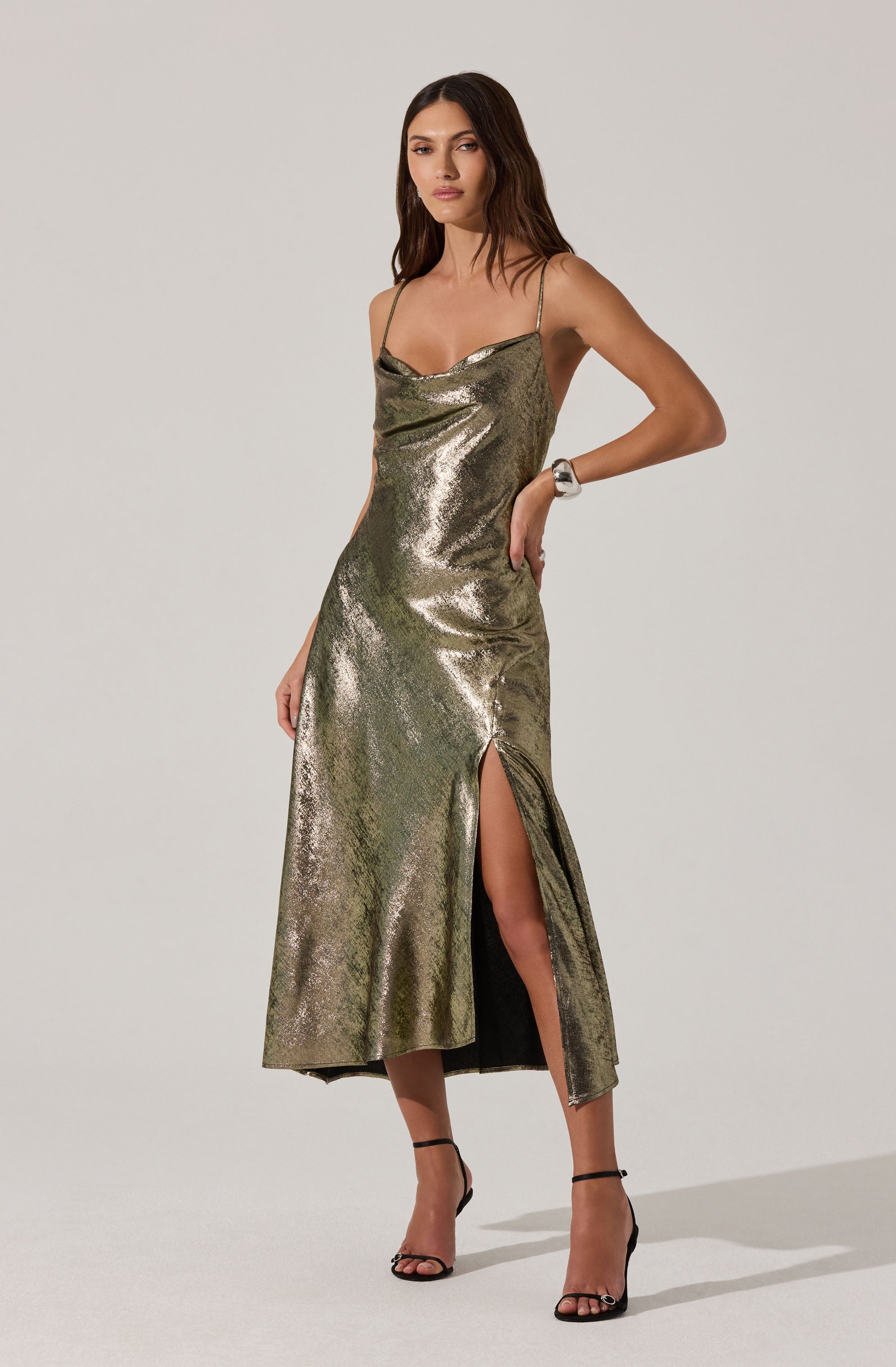 A woman is wearing a metallic, shiny, gold dress with spaghetti straps and a cowl neckline. The dress has a high slit on one side, revealing part of her leg. She is also wearing black high-heeled sandals with ankle straps.