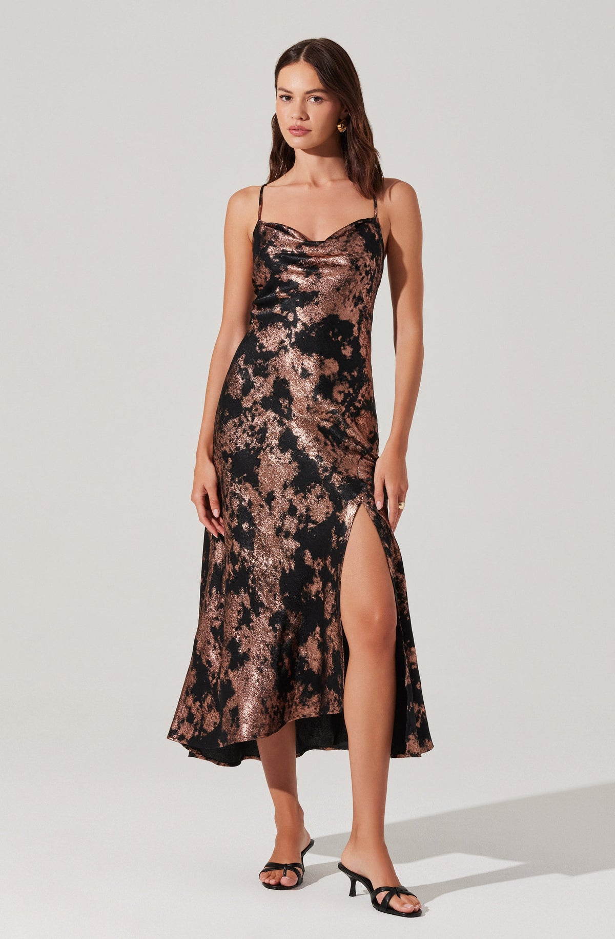 A woman is wearing a sleeveless, full-length slip dress with thin spaghetti straps. The dress has a metallic, abstract marbled pattern in shades of black and rose gold, with a high slit on one side that reaches above the knee. She is also wearing black high-heeled sandals with thin straps.