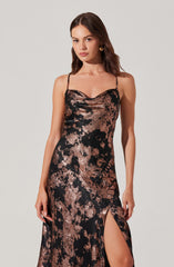 A woman is wearing a long, sleeveless dress with thin spaghetti straps. The dress features an abstract pattern in metallic copper and black, and has a high slit on one side.