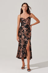 A woman is wearing a long, sleeveless dress with thin spaghetti straps. The dress has a shiny, metallic, abstract pattern in black and rose gold tones, with a high slit on one side. She pairs the dress with black open-toe heels and gold hoop earrings.