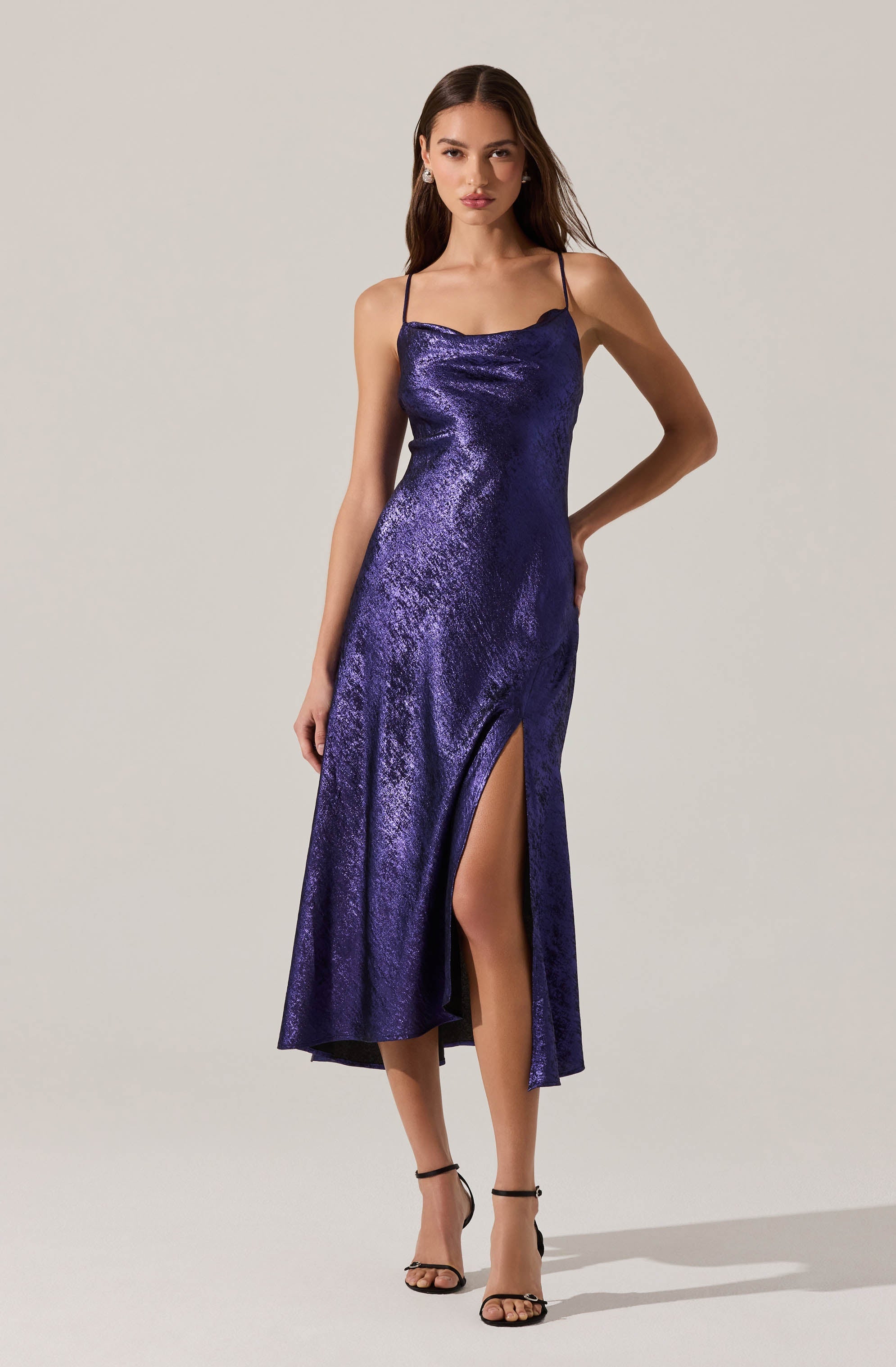 A woman is wearing a sleeveless, navy blue, shimmery slip dress with narrow spaghetti straps. The dress has a high slit on the left side that reveals her leg. She is also wearing black high-heeled sandals.