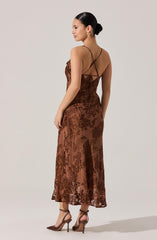 A woman is wearing a long, sheer brown lace dress with intricate floral patterns. The dress features thin spaghetti straps that crisscross at the back and tie at the mid-back. She is accessorizing with gold earrings and nude high-heeled shoes.