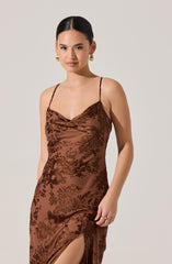 A woman is wearing a silky, brown slip dress with a floral pattern. The dress features thin spaghetti straps, a draped cowl neckline, and a high slit on the side. The fabric has a shiny, satin-like finish and the floral design is in a darker shade of brown.