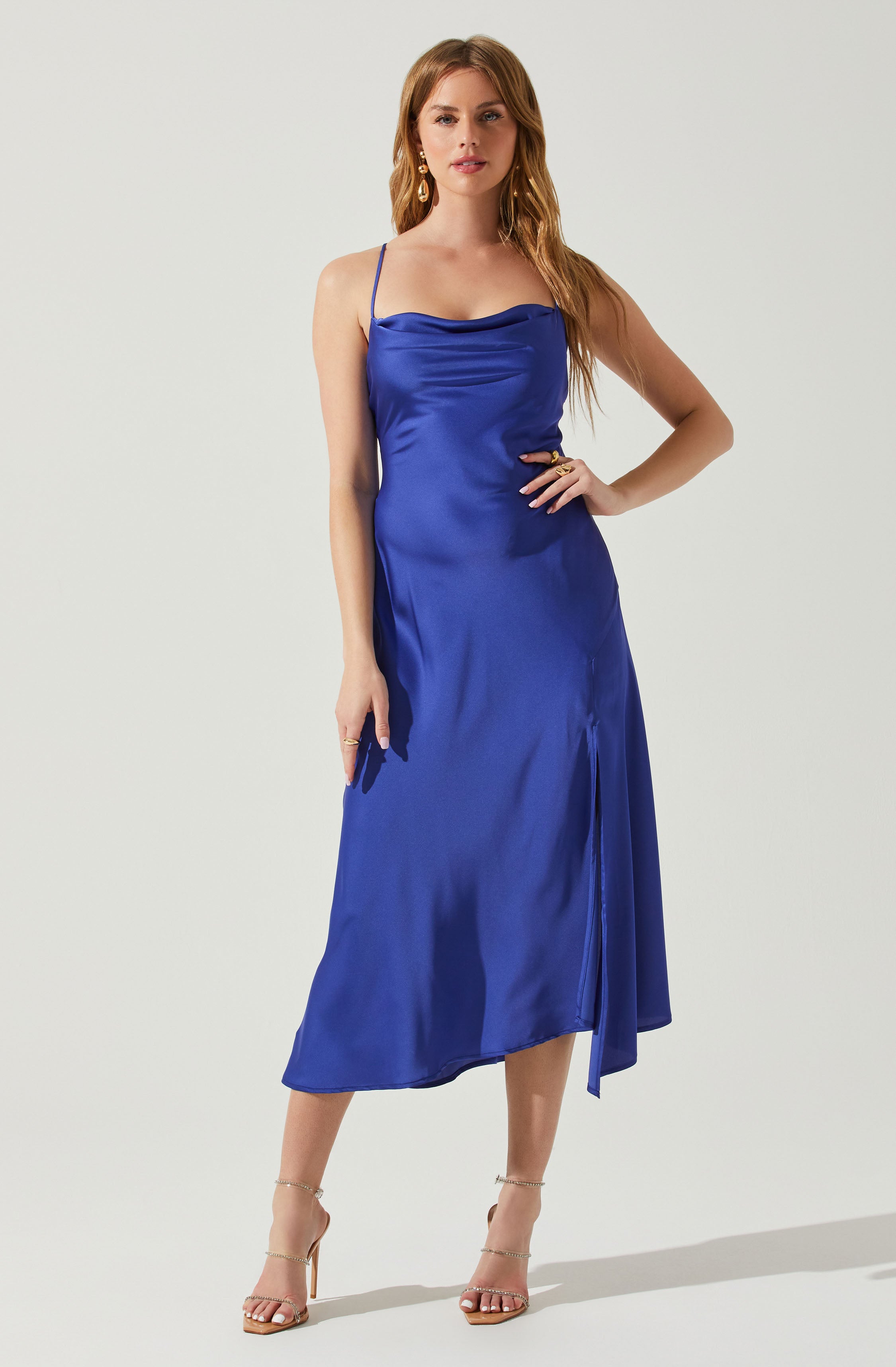 A woman is wearing a shiny, royal blue, satin slip dress with thin spaghetti straps. The dress features a cowl neckline and a slit on the front. She accessorizes with dangling earrings, rings, and nude high-heeled sandals with ankle straps.