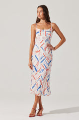 A woman is wearing a sleeveless, spaghetti strap midi dress with a white background and a colorful print of various words and phrases in red and blue. She has paired the dress with red high-heeled sandals.
