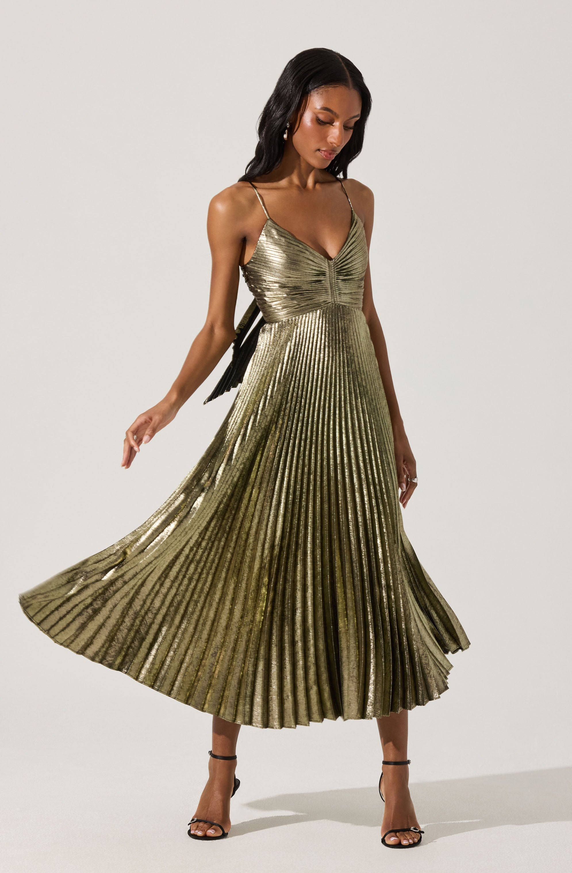 A woman is wearing a sleeveless, metallic gold dress with thin spaghetti straps. The dress features a fitted bodice with a gathered detail at the center and a flowing, pleated skirt that reaches mid-calf length. She pairs the dress with black high-heeled sandals.
