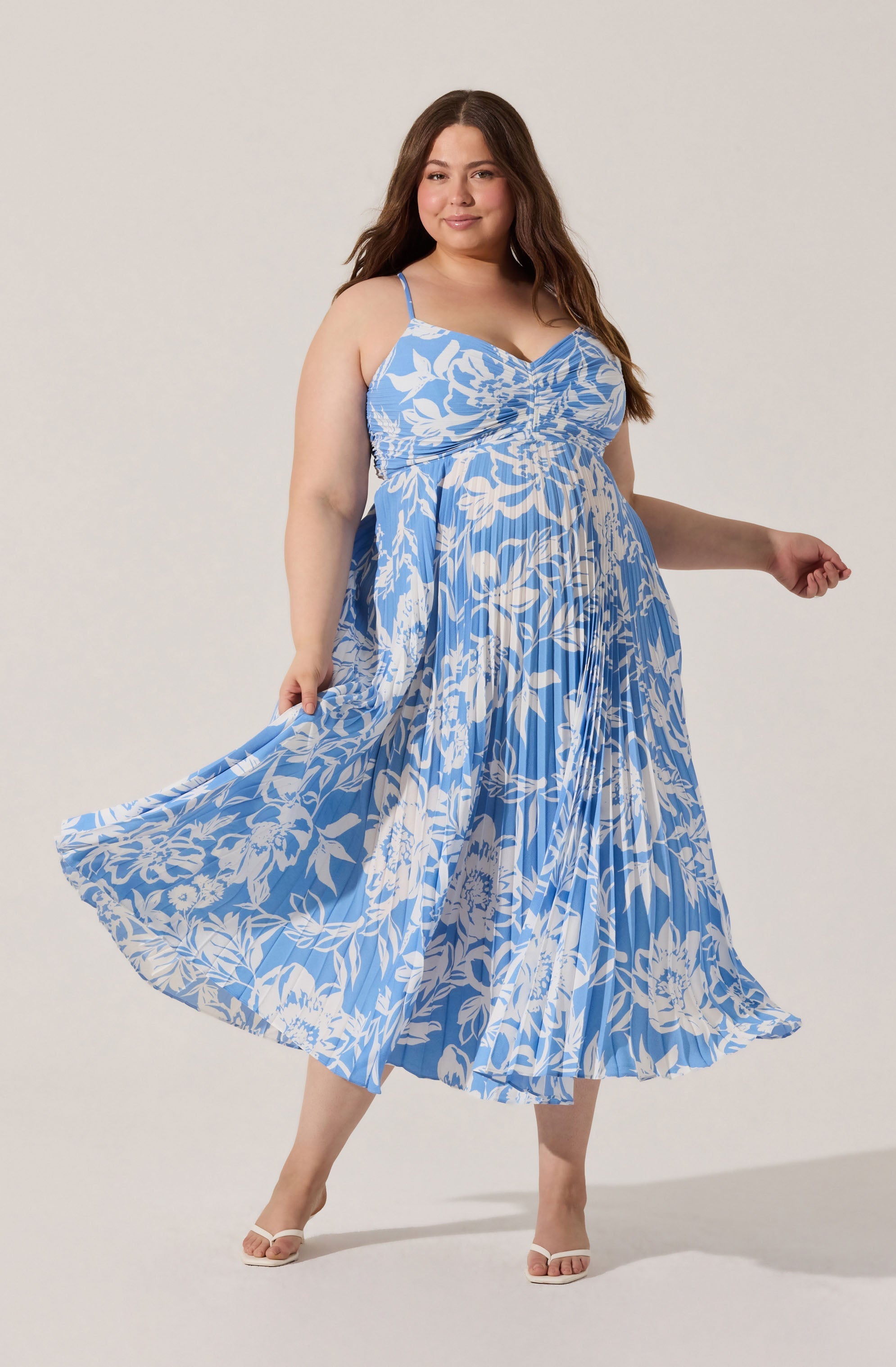 A woman is wearing a sleeveless, floral-patterned dress with thin spaghetti straps. The dress features a sweetheart neckline with gathered fabric at the bust, and the skirt has pleats with a blue background and white floral design. The fabric appears lightweight and flowing. She is also wearing light-colored, open-toe sandals.