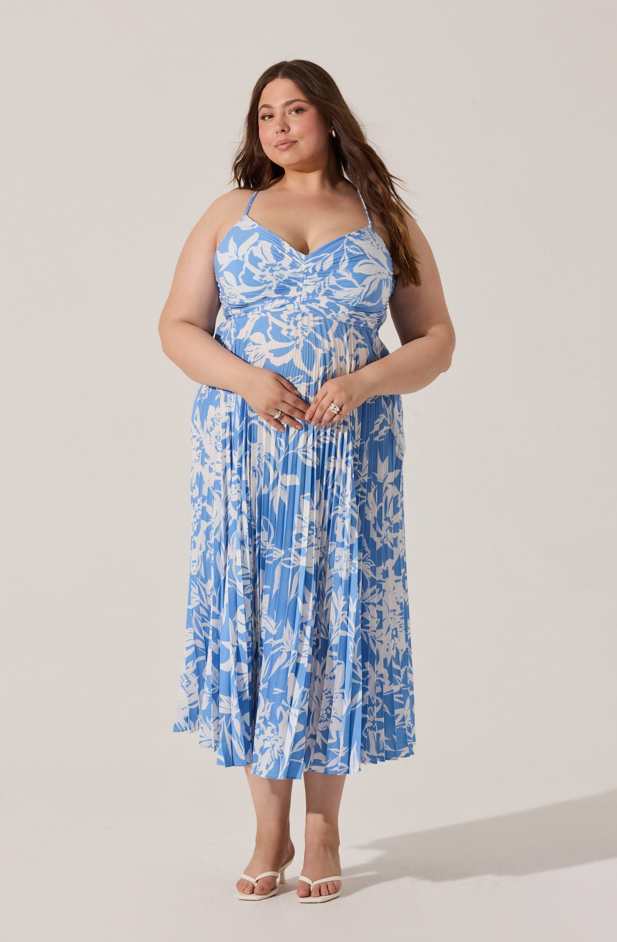 A woman is wearing a long, flowy, sleeveless dress with thin spaghetti straps. The dress features a blue and white botanical floral pattern and has a gathered detail at the bust. She is also wearing white high-heeled sandals.