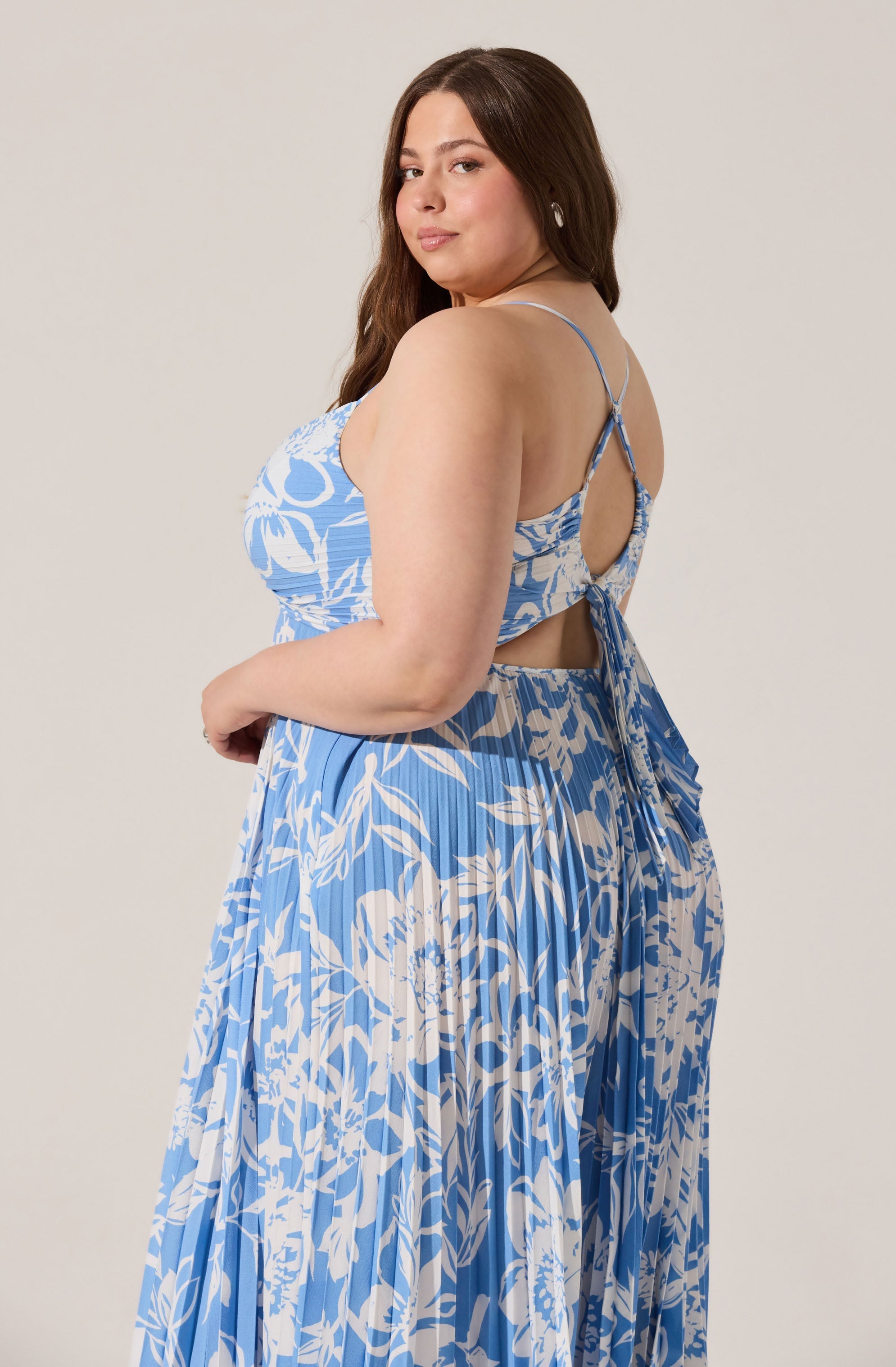 The image features a woman wearing a matching blue and white floral outfit. The top has thin spaghetti straps and a back cutout tied with a bow, showing a portion of her back. The skirt has a high waist and is pleated, with a flowing, maxi length. Both the top and skirt display an abstract floral pattern in shades of blue and white.