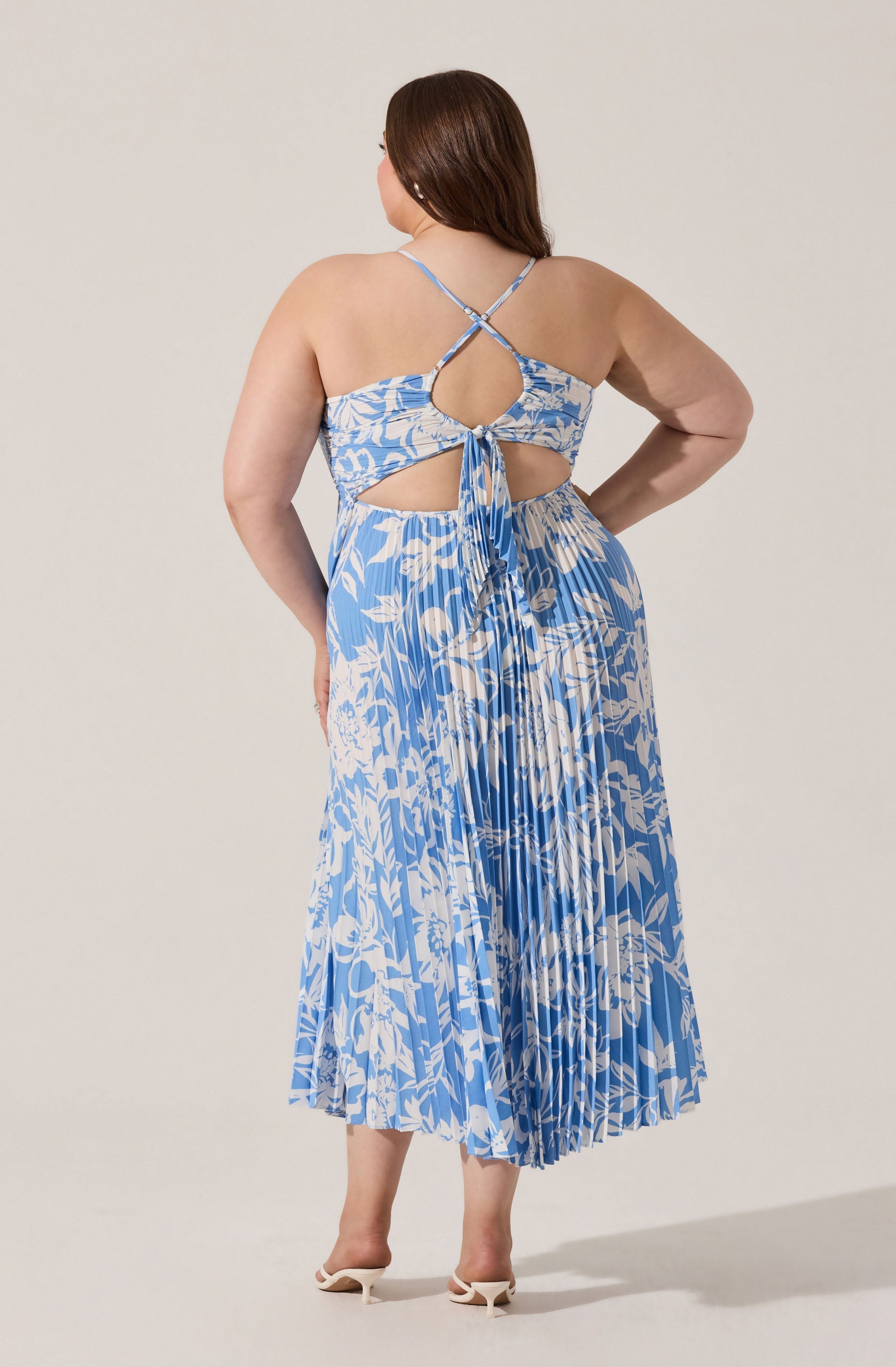 A woman is wearing a sleeveless, blue and white floral maxi dress with pleats. The dress features a cut-out design at the back with a fabric tie and crisscross spaghetti straps. She is also wearing a pair of white high-heeled shoes.