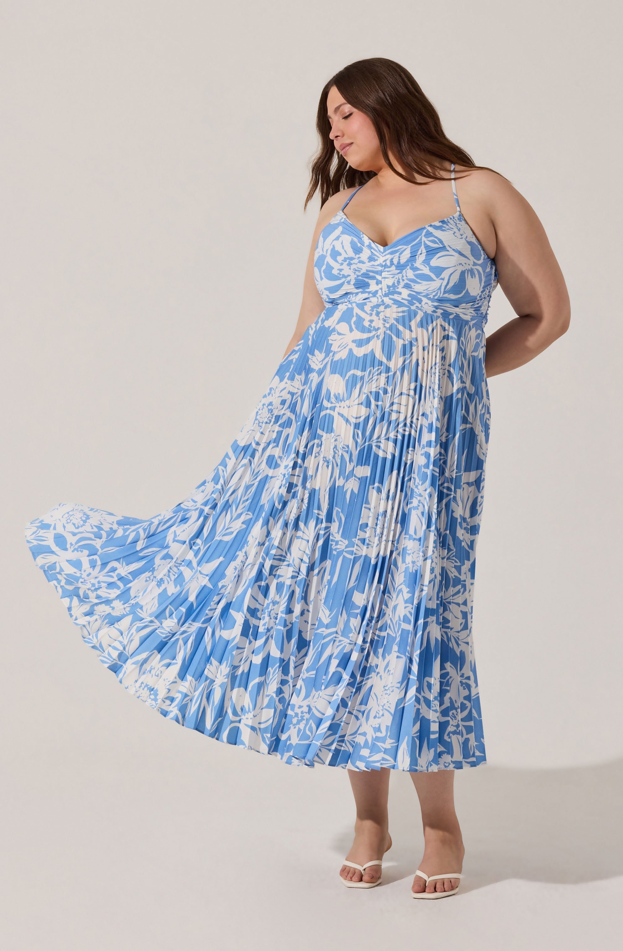 A woman is wearing a sleeveless, ankle-length dress with spaghetti straps. The dress is light blue with a white floral pattern. She is also wearing white open-toe sandals.