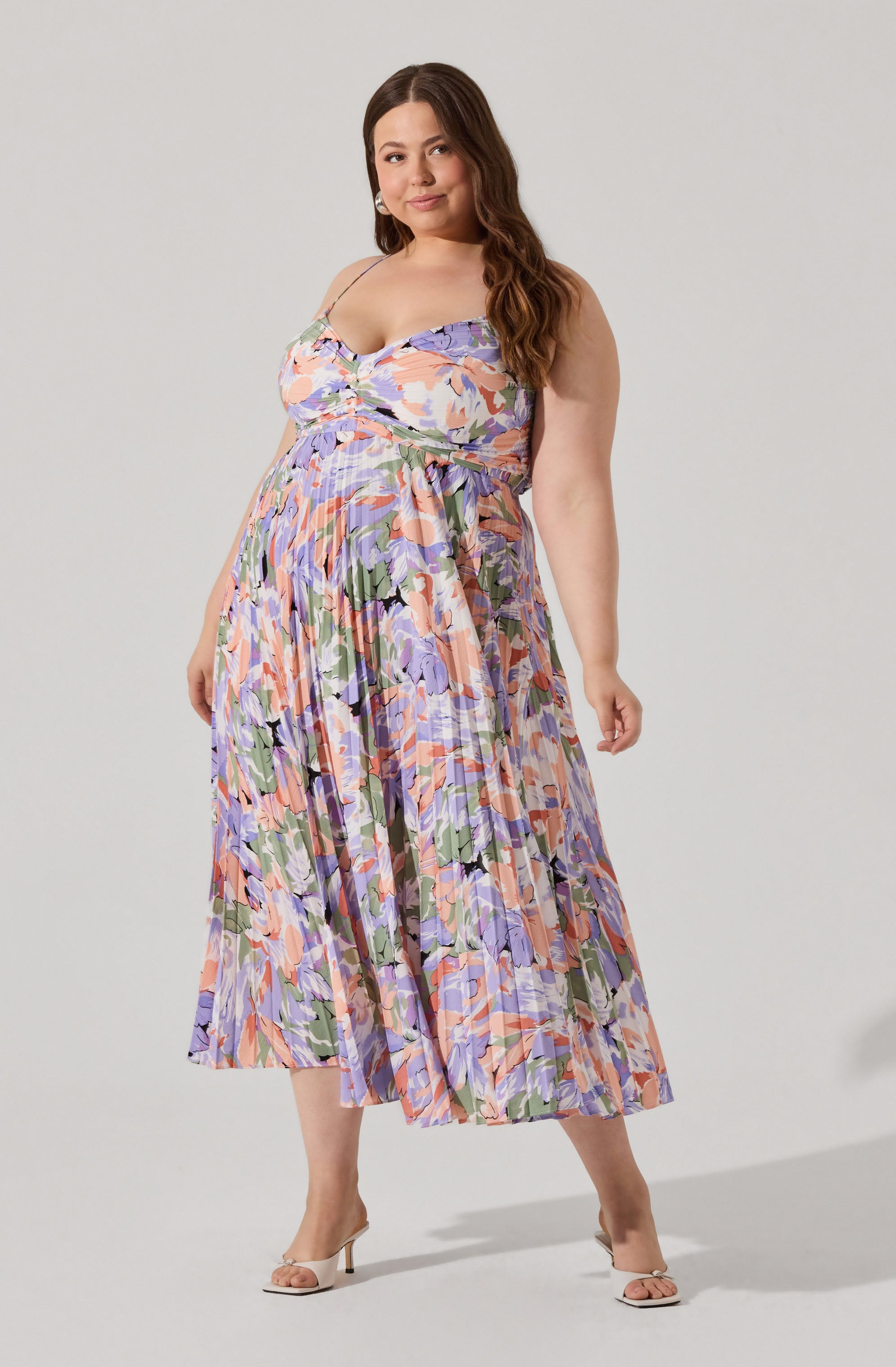 A woman wearing a sleeveless, floral-patterned dress with spaghetti straps. The dress features a mix of pastel colors including purple, pink, peach, and green in an abstract floral design. She is also wearing white high-heeled sandals.